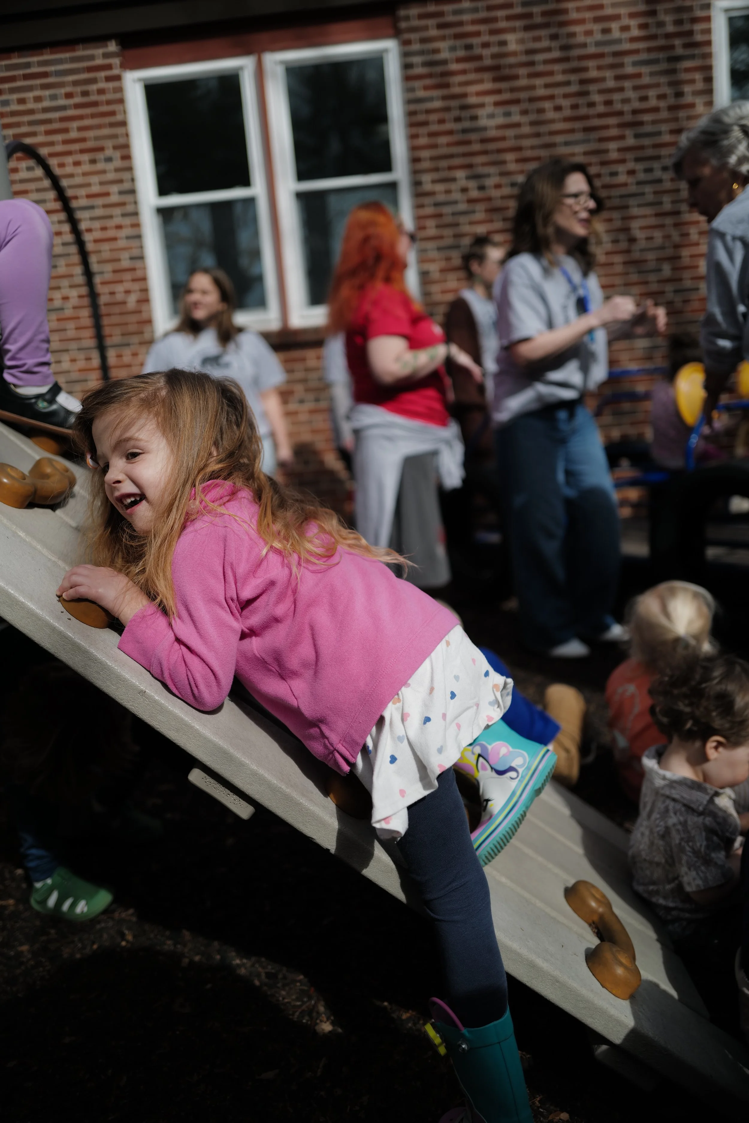 A young girl climbing playfully on a slide at a playground, with a group of adults and children in the background near a brick building.