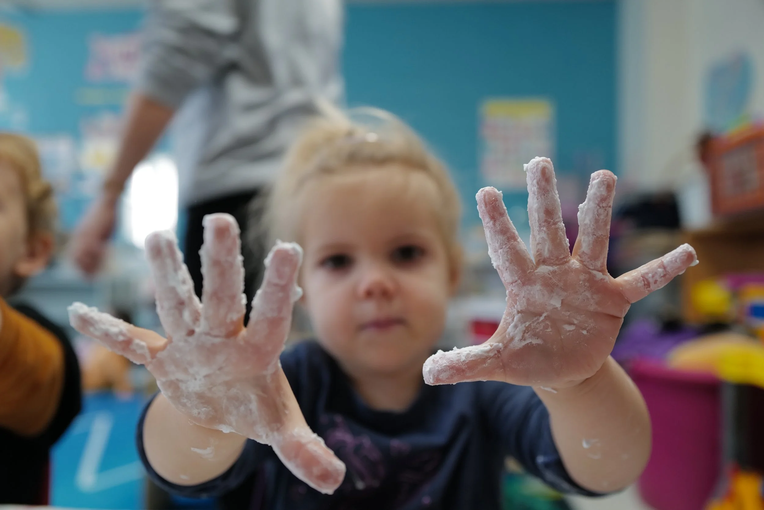 A young girl with shaving cream-covered hands reaching towards the camera in a classroom setting.