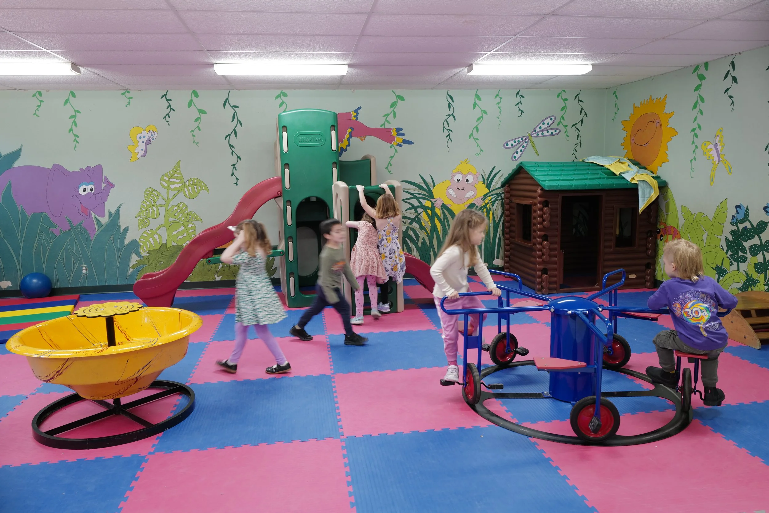 Children playing in an indoor playground with colorful mats, a slide, a house-shaped play structure, and various riding toys. The walls are decorated with cartoon jungle animals and plants.