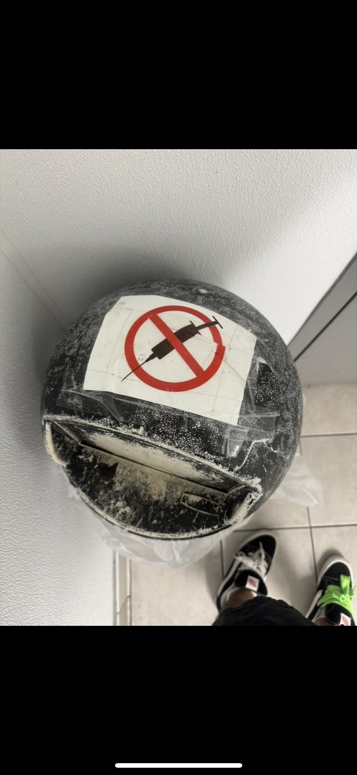 Dusty plastic helmet with a no syringes sign on top, viewed from above, on a gray surface next to a person's legs and shoes.