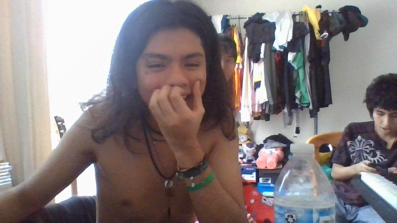 A shirtless young person with dark, wavy hair is smiling and resting their chin on their hand. They are inside a room with a wardrobe in the background filled with hanging clothes and various items on shelves. There is another person sitting nearby, looking down at a keyboard, in front of a water bottle and other items.