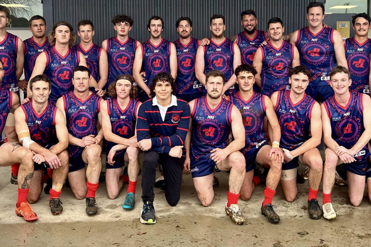 A group of young men posing for a team photo in sports jerseys and shorts, some kneeling and some standing, against a metal building background.