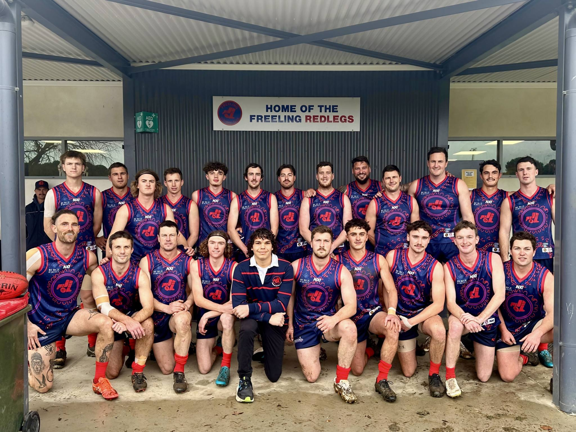 A group of 19 Australian football players posing together under a shelter at the home of the Freeling Redlegs. They are wearing blue and red team uniforms, some are kneeling in front, and others are standing behind, smiling at the camera.