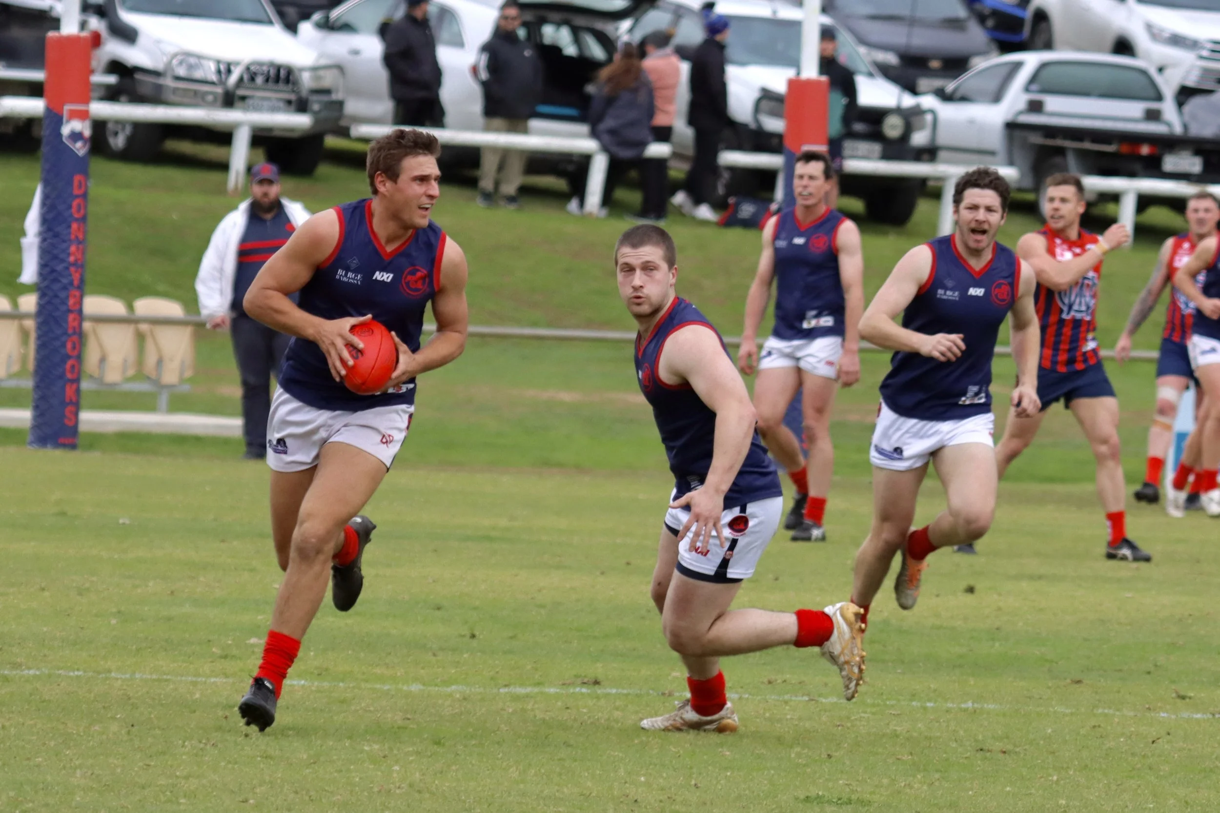 Australian rules football players in navy and white uniforms in action on a grassy field, with spectators and parked cars in the background.