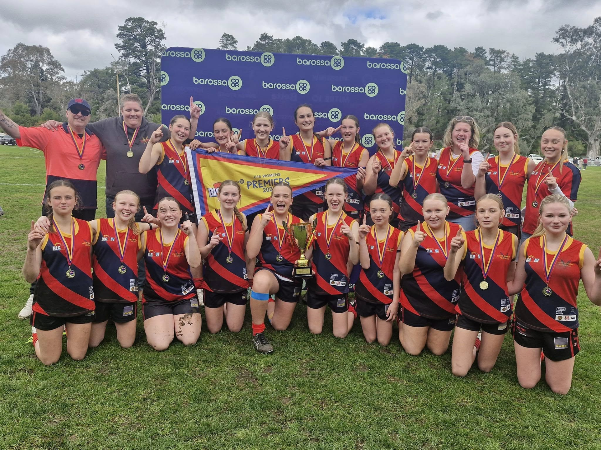 A girls' sports team celebrating their victory, holding a banner that says 'Premiers 2023,' with medals around their necks, a trophy, and posing on a grassy field.