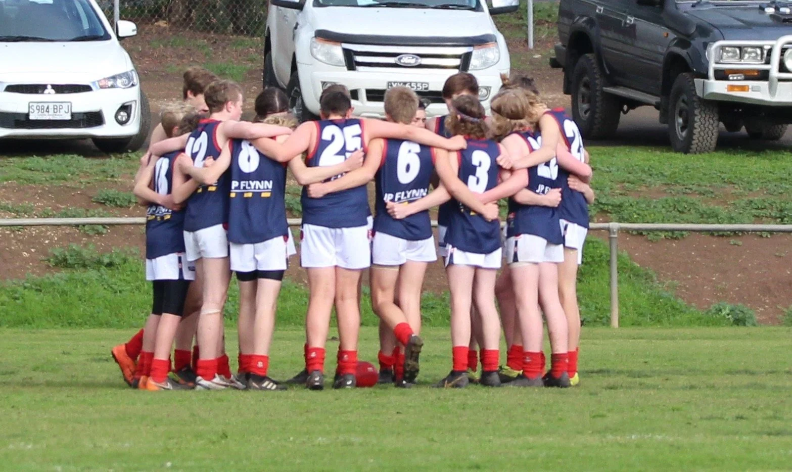 A group of young female athletes in blue and white sports uniforms with red socks huddled together with arms around each other on a grassy field, preparing for a game.