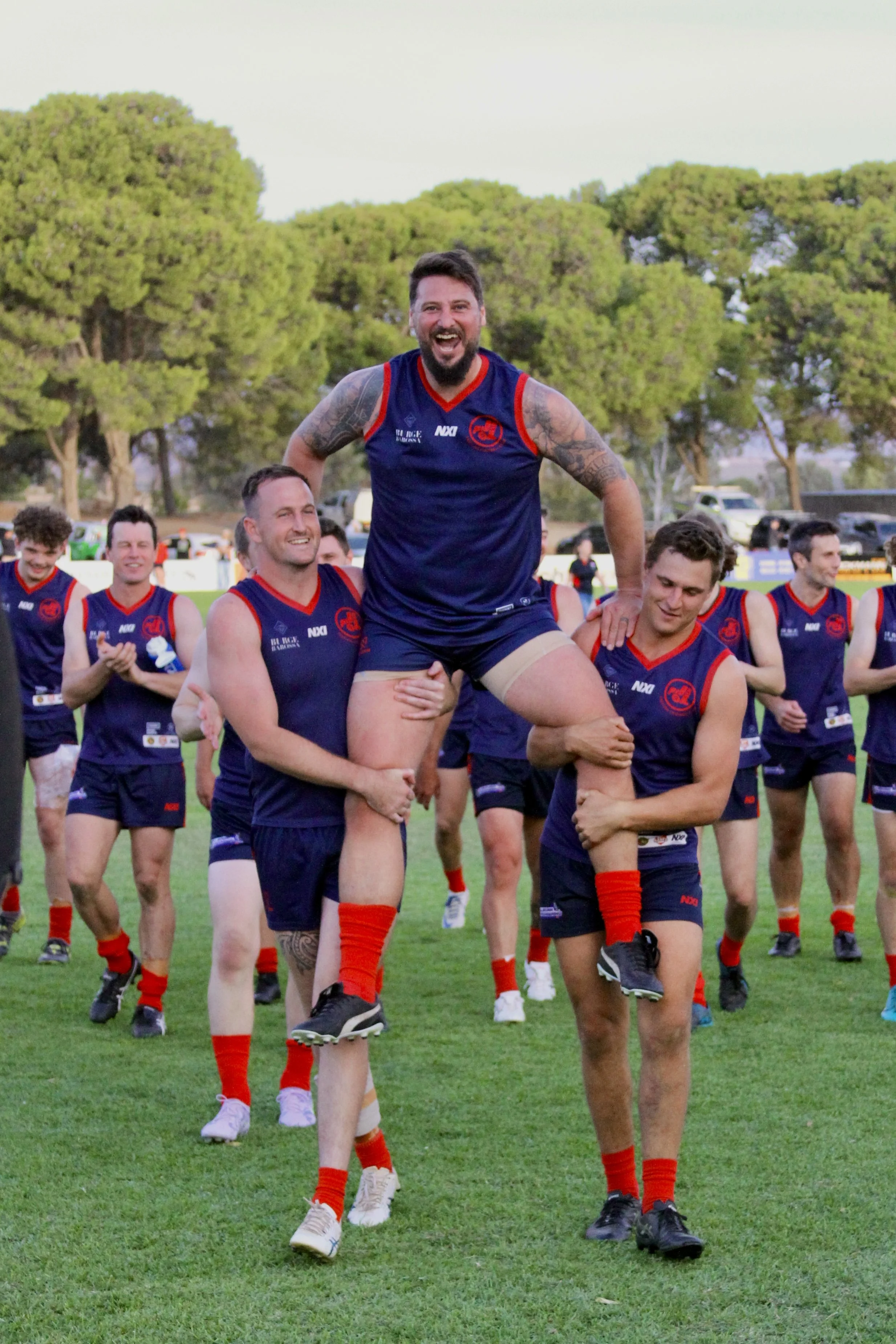 A group of Freeling footballers in navy blue and red sports uniforms celebrating on a grassy field, with one player being lifted by two others.
