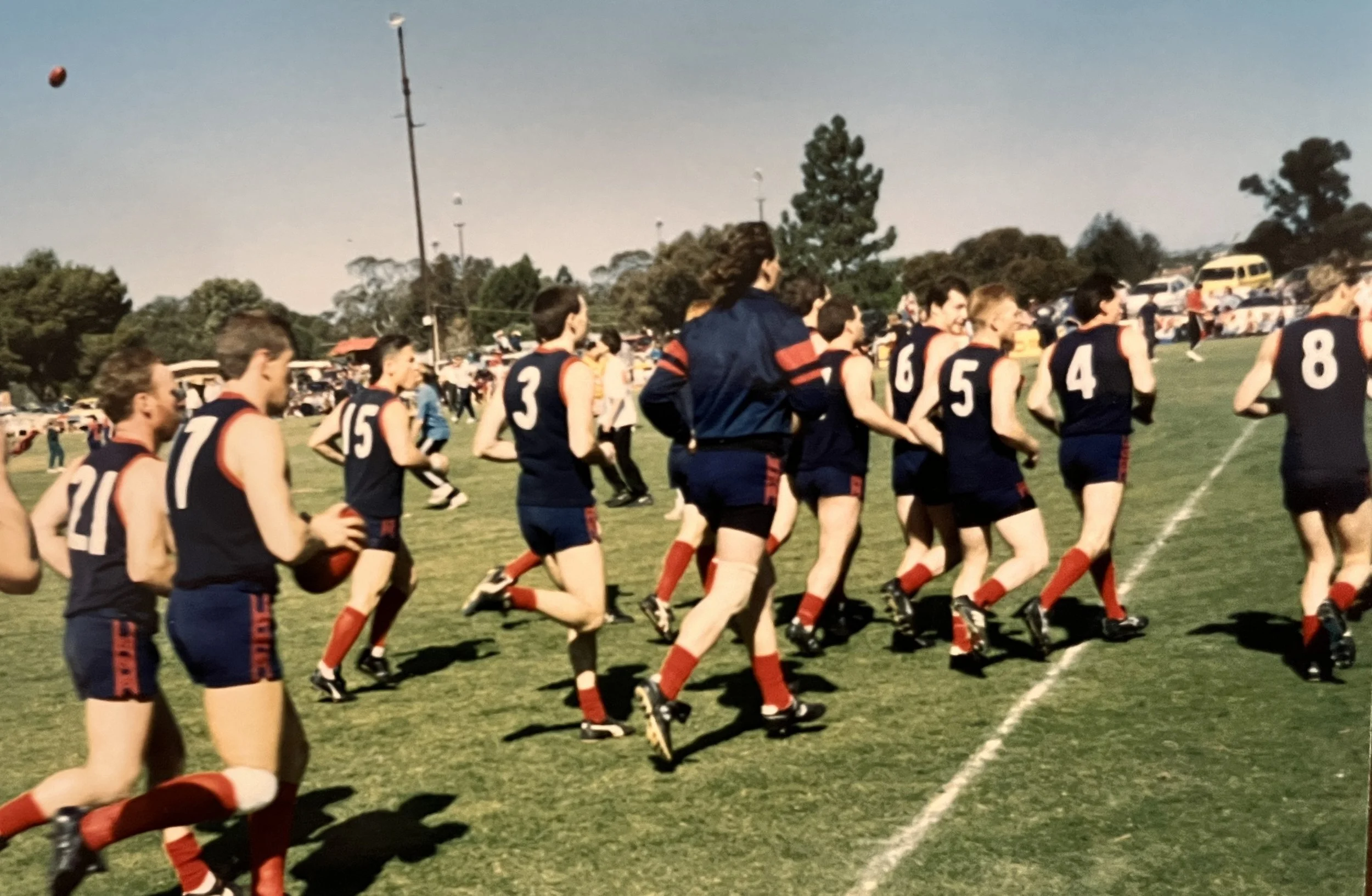 A group of young men running on a grassy sports field, wearing navy blue uniforms with red accents and numbering, with a line of trees and parked cars in the background.