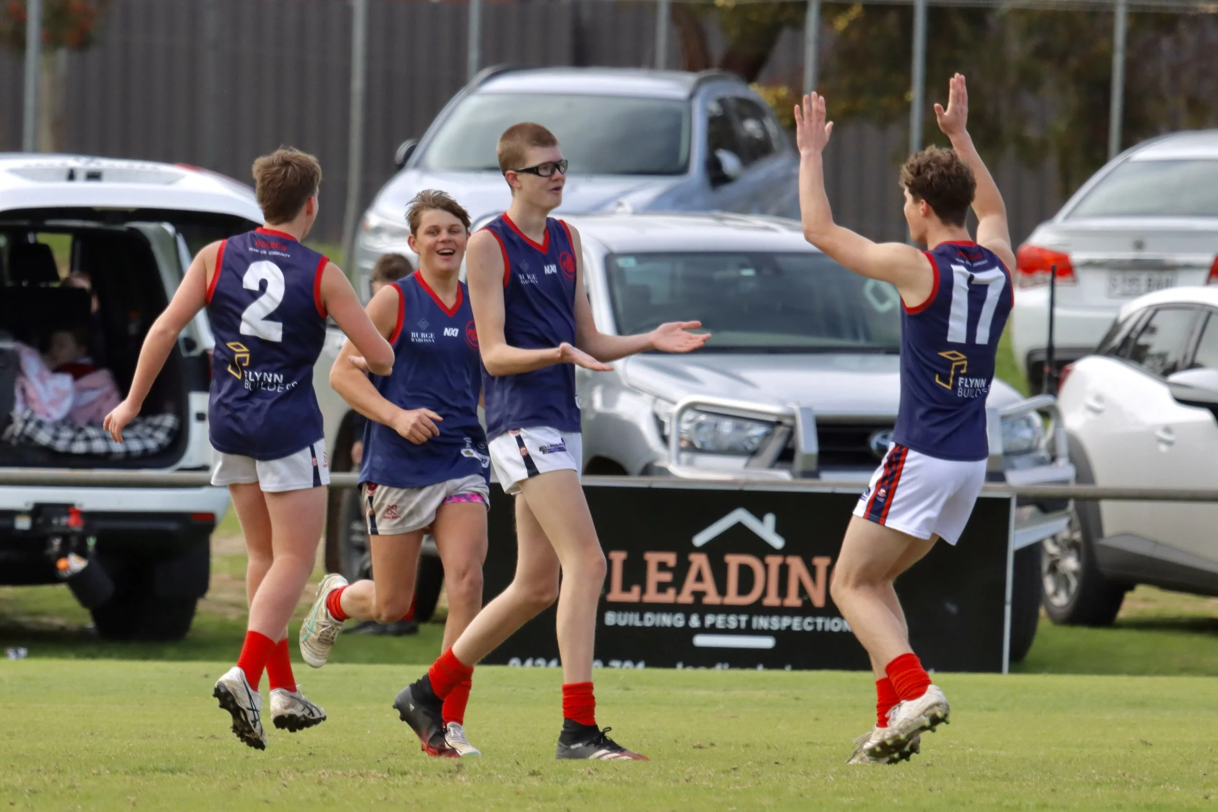 Freeling footballers in blue and red sports uniforms celebrating on a grassy field during a daytime game, with parked cars and a building in the background.
