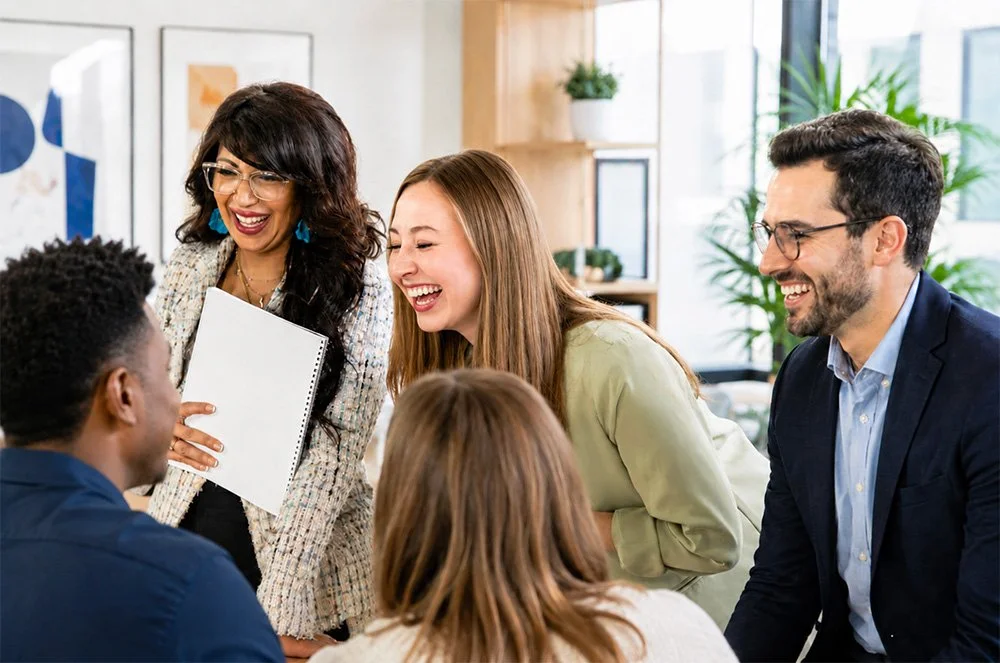 Five diverse colleagues in a bright office space, laughing and smiling during business training in Redding, CA led by Sarah Bliss Daly