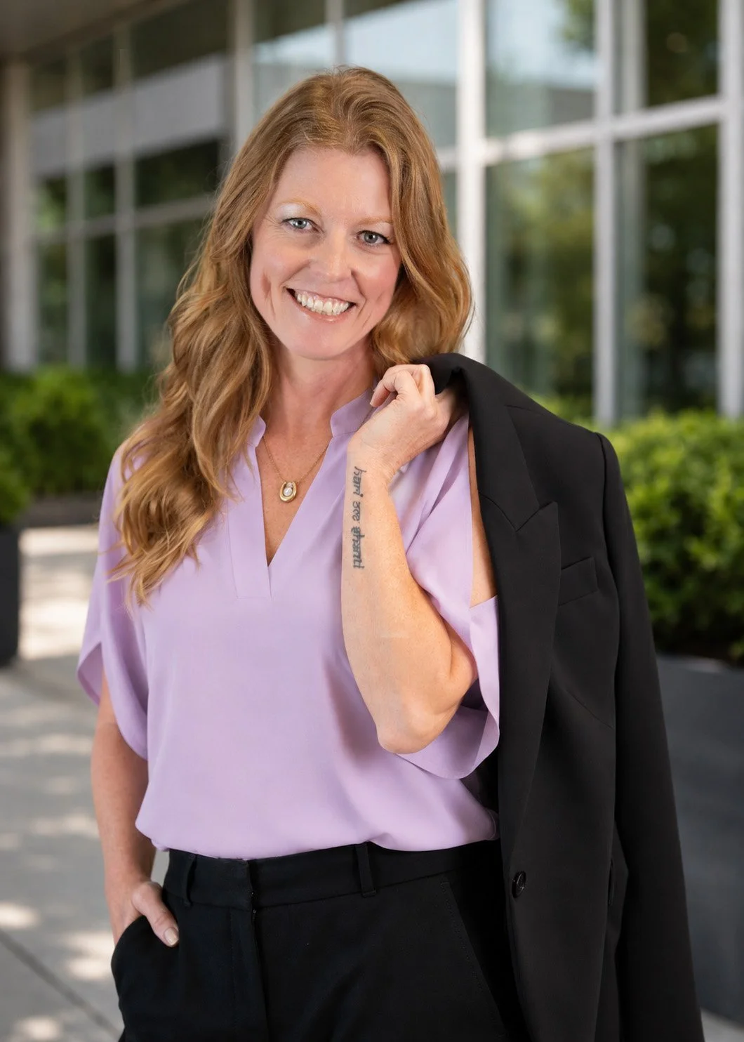 Sarah Bliss Daly, personal coach and keynote speaker in Redding, CA wearing a lavender blouse, black pants, and holding a black blazer over her shoulder