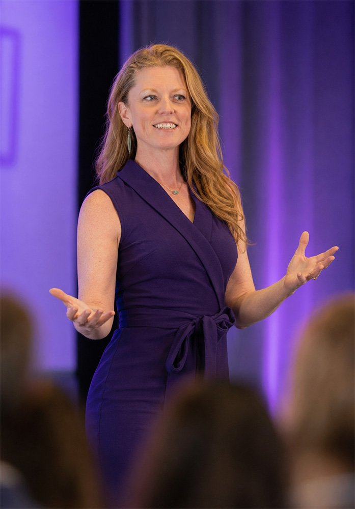 Sarah Bliss Daly, keynote speaker in Redding, CA wearing sleeveless purple dress is speaking to an audience in a conference room with purple lighting.
