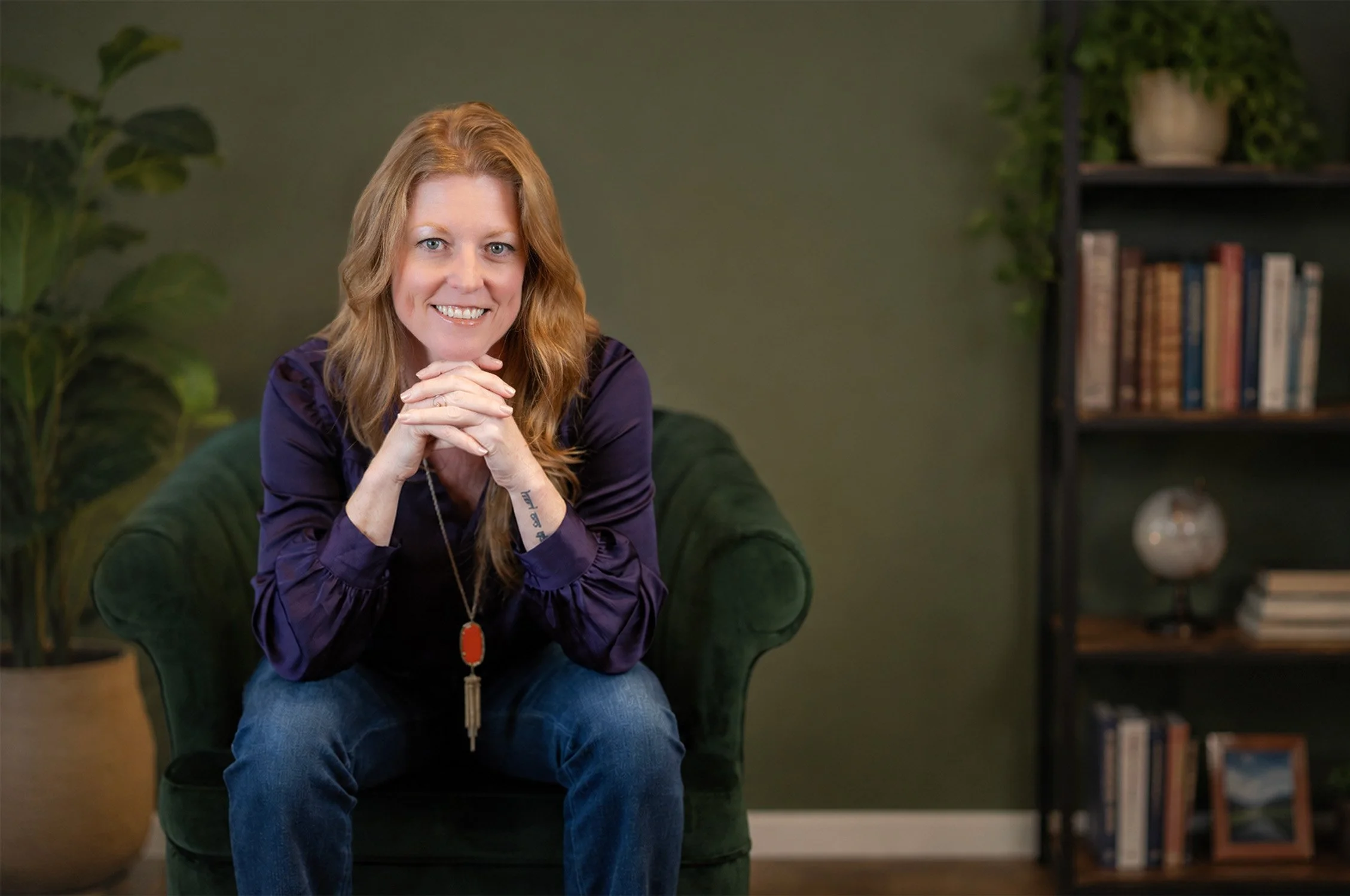 Sarah Bliss Daly, personal coach and keynote speaker on a green armchair, smiling at the camera, in a room with a dark green wall, a bookshelf with books, and plants.