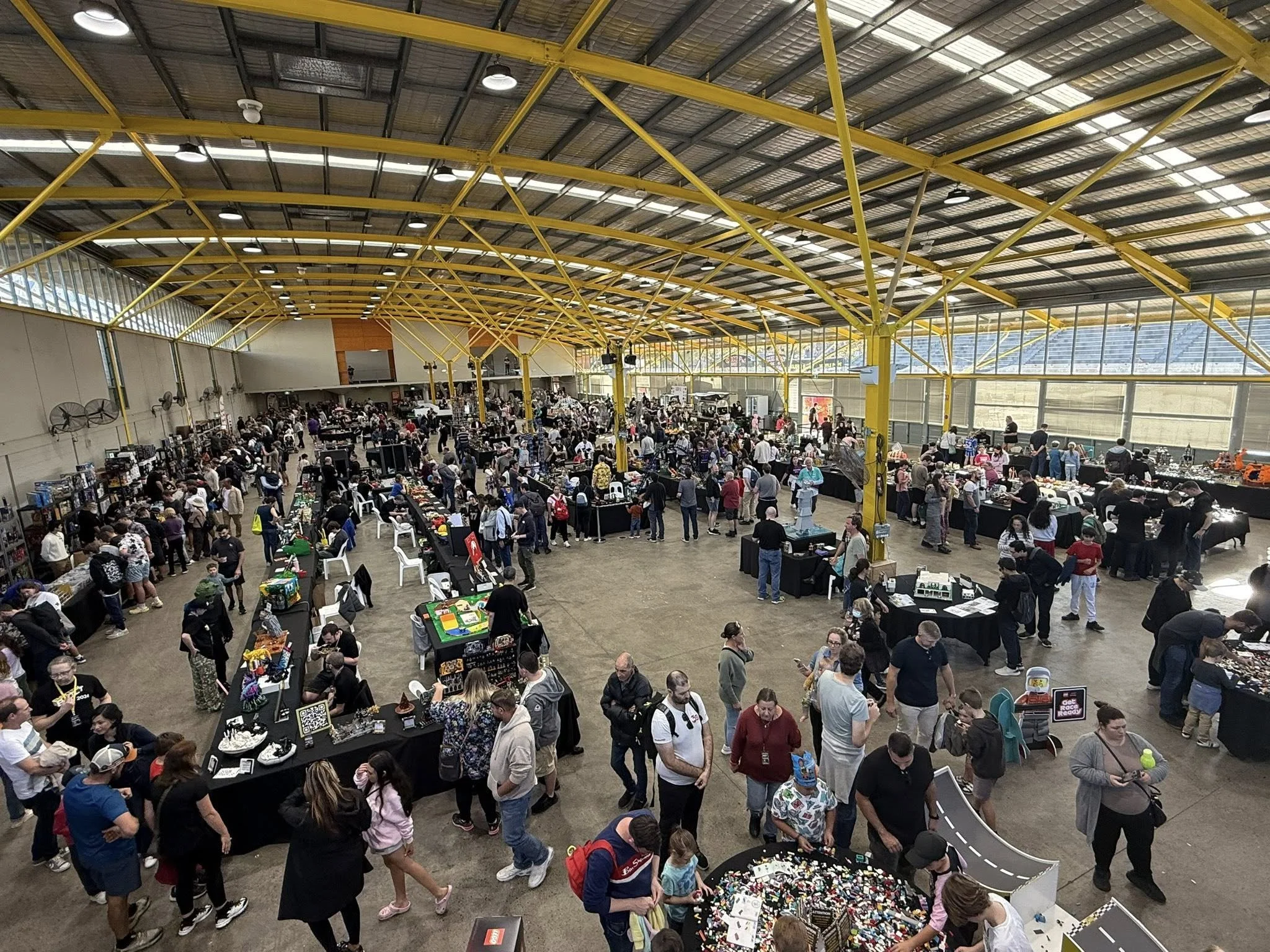 Crowd of people shopping at a toy and collectibles fair inside a large industrial-style building with yellow steel beams and glass windows