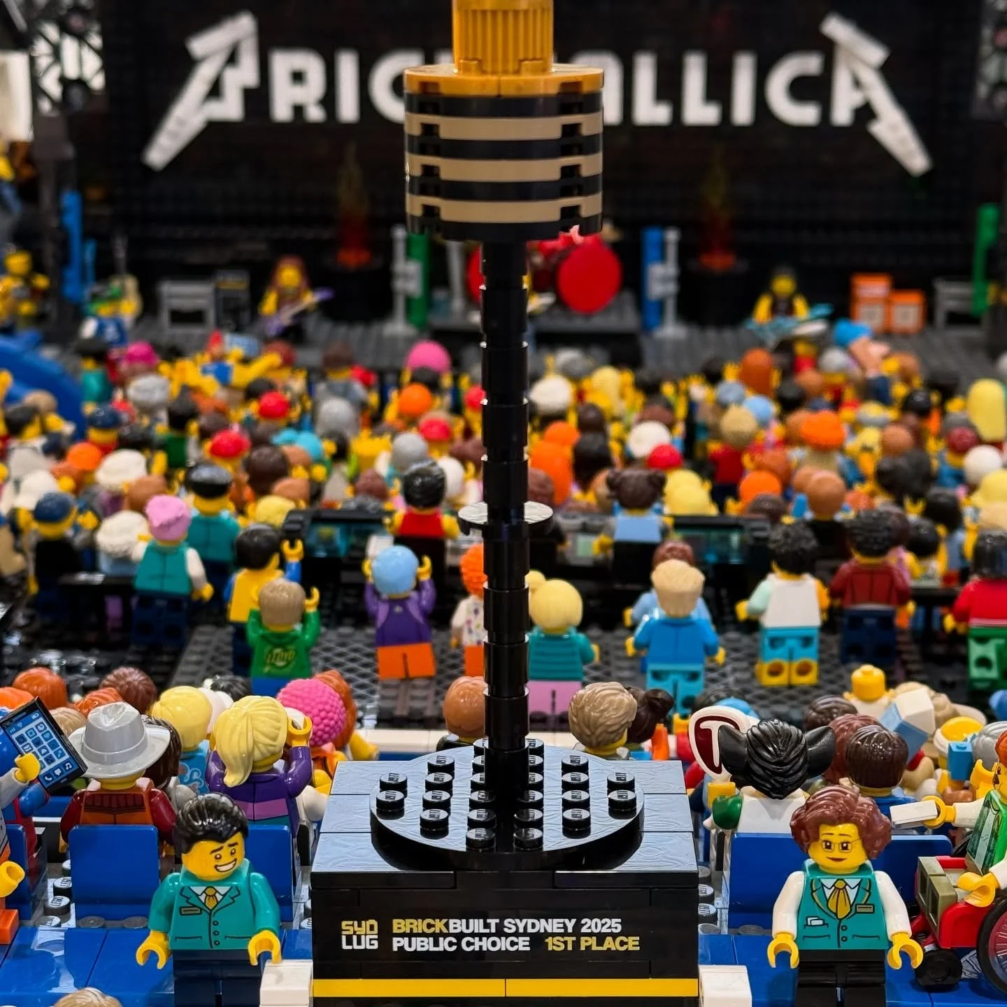 Close-up of a LEGO display at the BrickBuilt Sydney 2025 event, featuring a large microphone and a sign indicating first place in a public choice competition with numerous LEGO minifigures and a stage in the background.