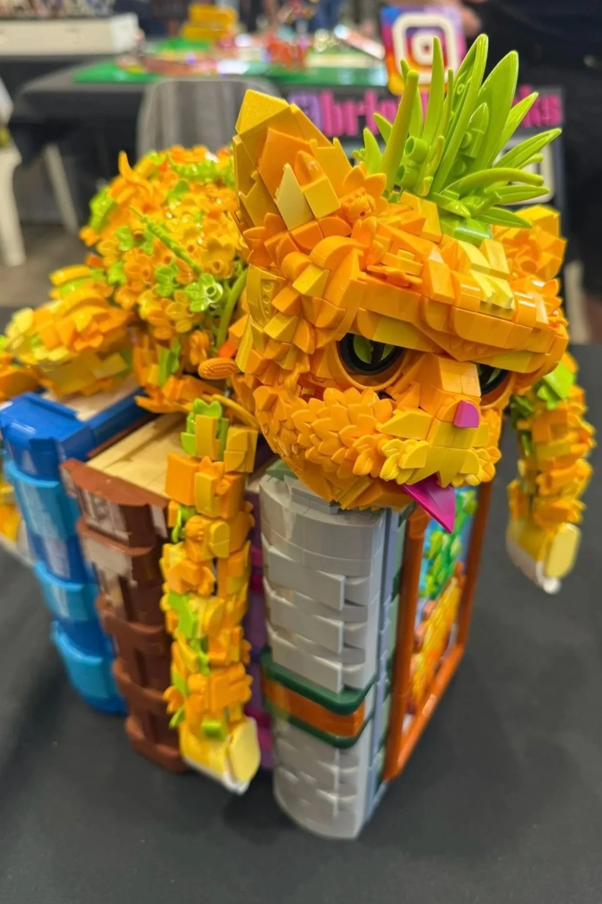 Colorful LEGO sculpture of a lion with a mane, perched on a cube-shaped display with various LEGO bricks around it.