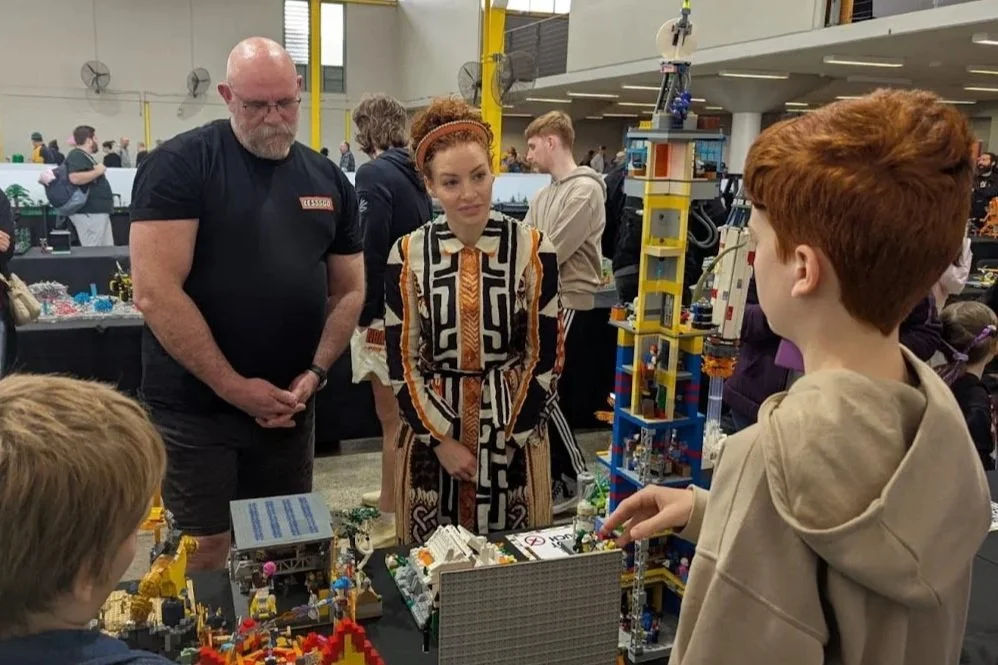 A LEGO building display at an event where a young boy is explaining the LEGO structure to a woman and a man. Other people are in the background browsing booths.