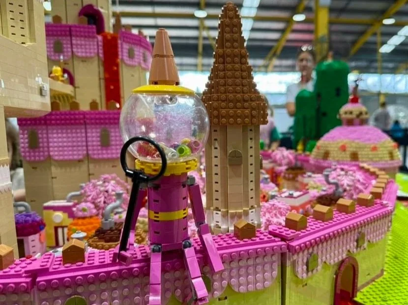 A LEGO castle scene with pink and beige structures, a pink tower, and a clear bubble with toys on top, set on a green base. In the background, people are observing.