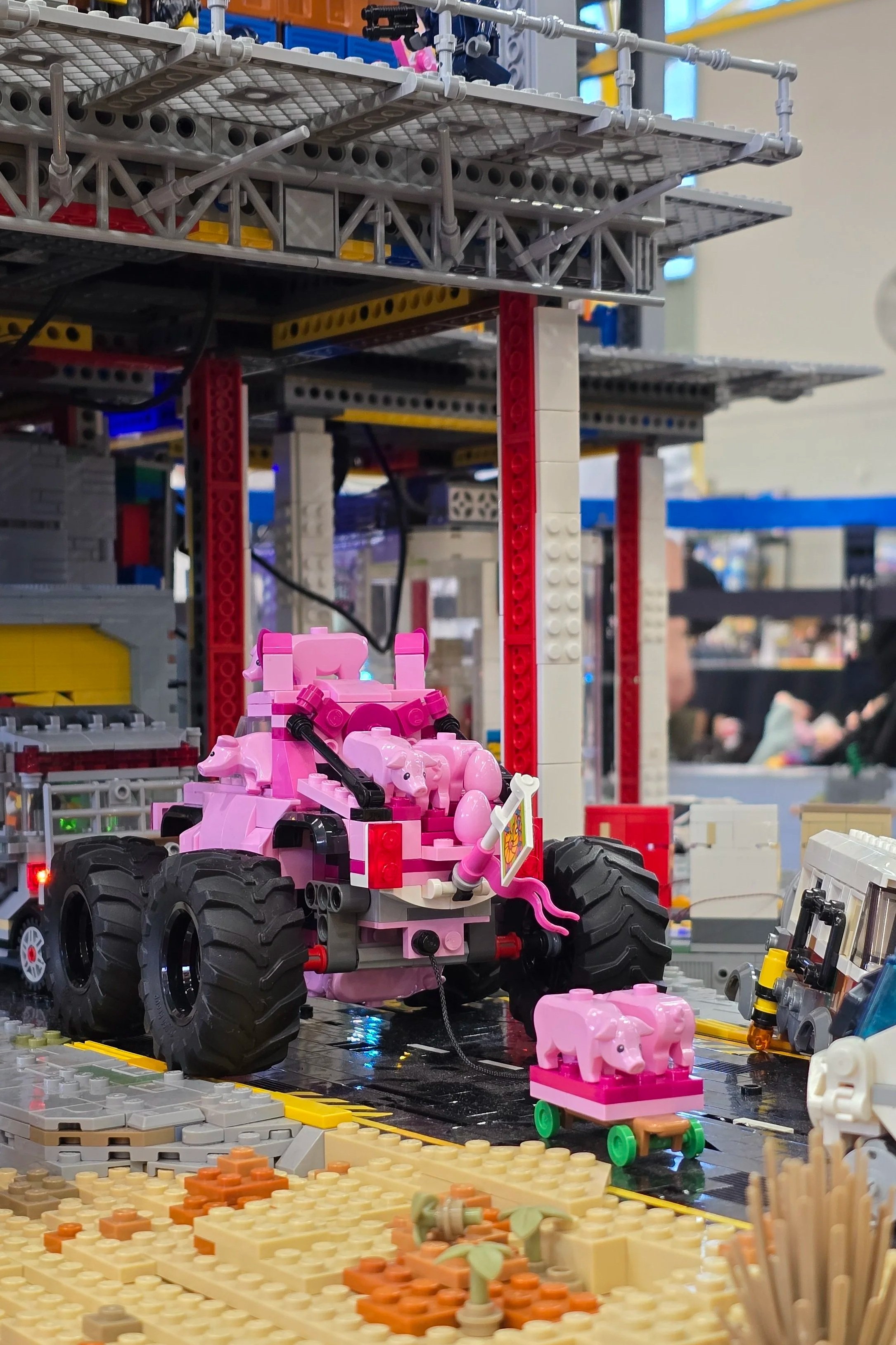 A LEGO display of a pink monster truck with a pig figure attached to a cart and small pig figures on its back, set on a black platform with construction and building elements around it.