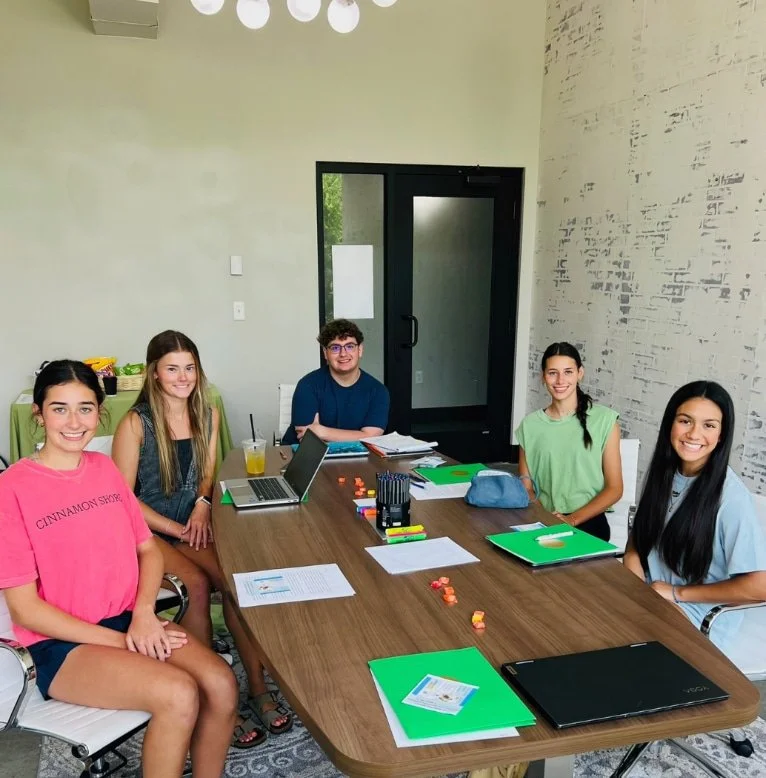 Five young adults sit around a table with laptops and folders, smiling as they work on college prep in a bright meeting room.