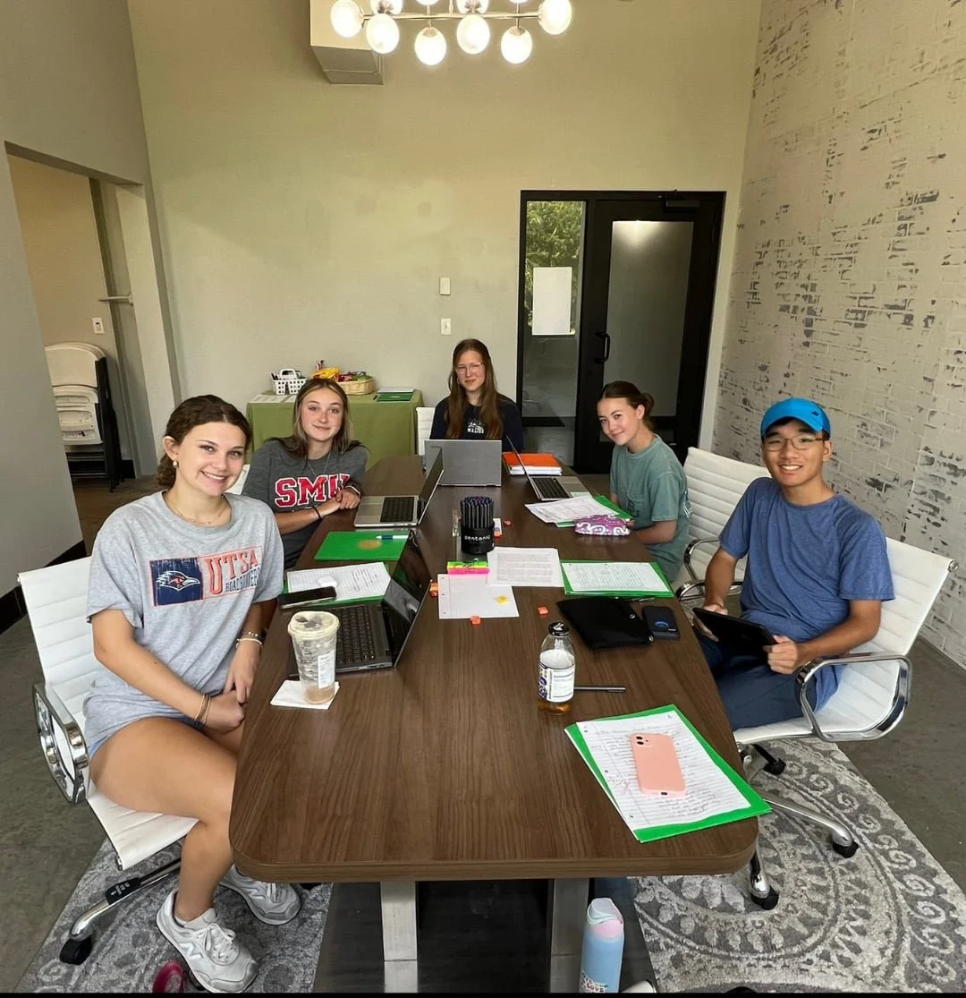 Five high school students sit around a table with laptops, smiling as they work on college prep in a bright, modern meeting room.