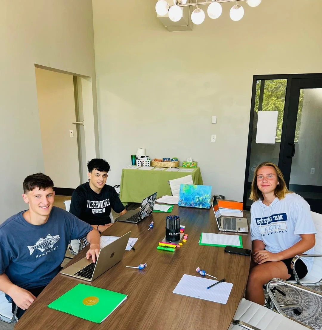 Three students sit at a table with laptops and notebooks, focused on college prep in a bright, modern study room.