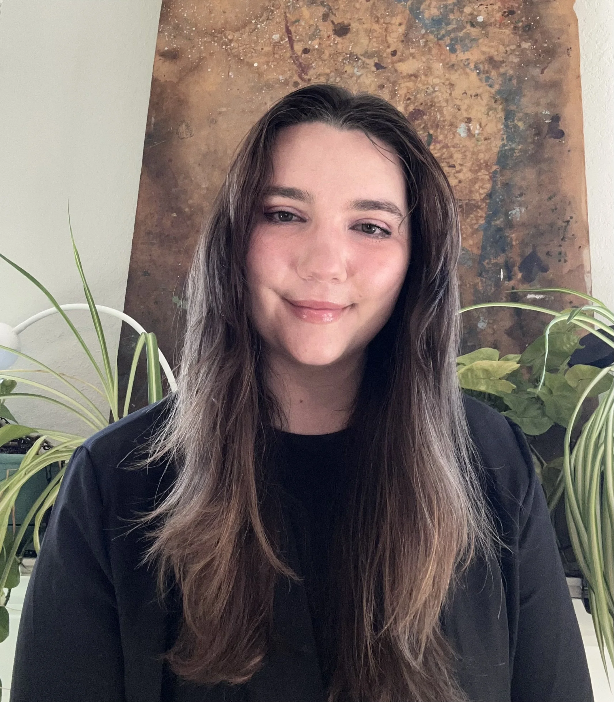 A woman with long brown hair and light skin smiling in front of a textured abstract art piece with earthy tones, surrounded by green houseplants.