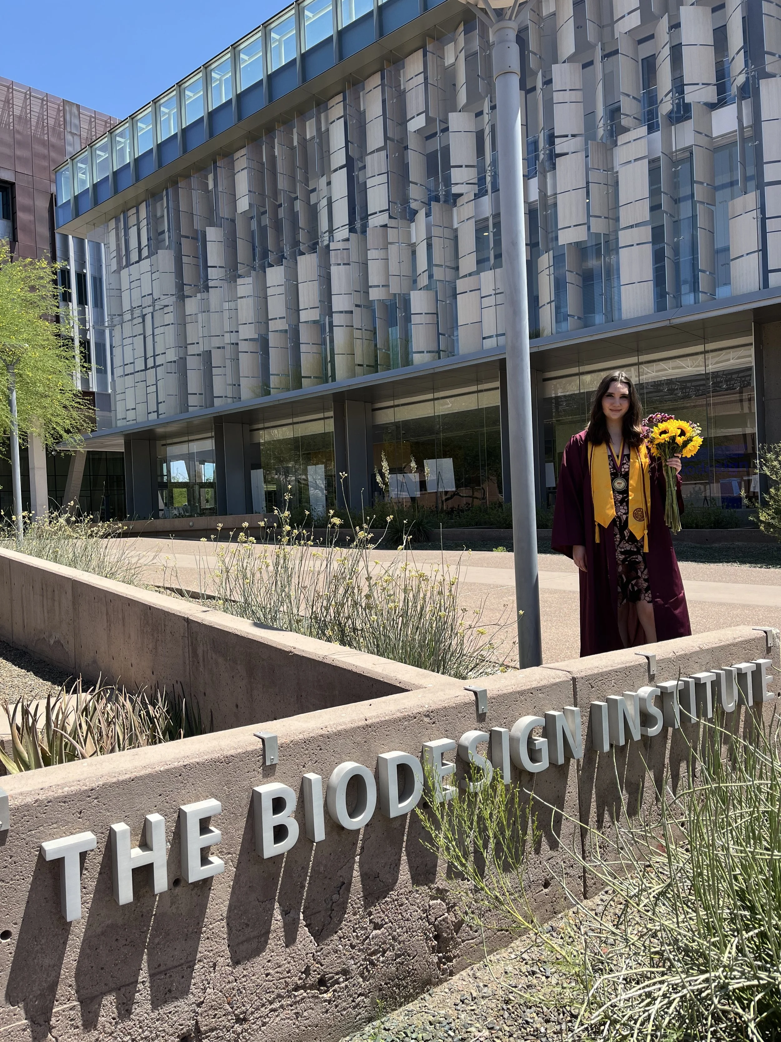 A young woman in graduation attire, holding a bouquet of sunflowers, standing in front of a modern building with glass windows and a concrete sign that reads "THE BIODESIGN INSTITUTE".