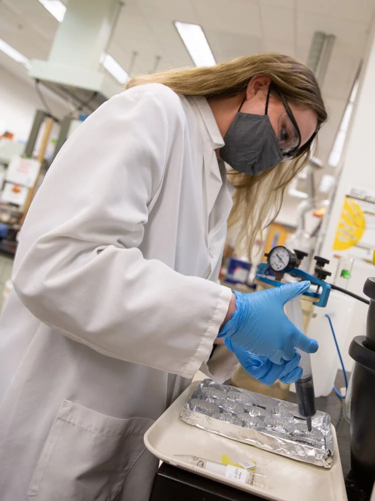 A woman scientist wearing a face mask, glasses, a white lab coat, and blue gloves working with a pipette in a laboratory.