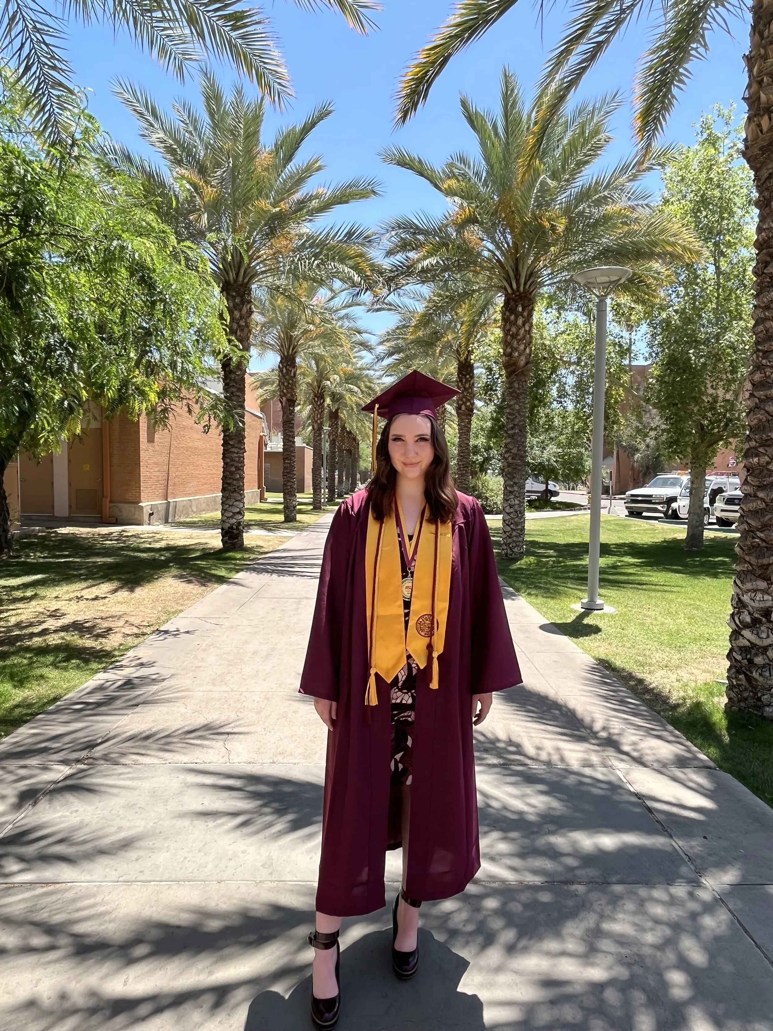 A woman in a maroon graduation gown and cap, with a gold stole, standing on a pathway lined with palm trees, smiling during her graduation celebration on a sunny day.