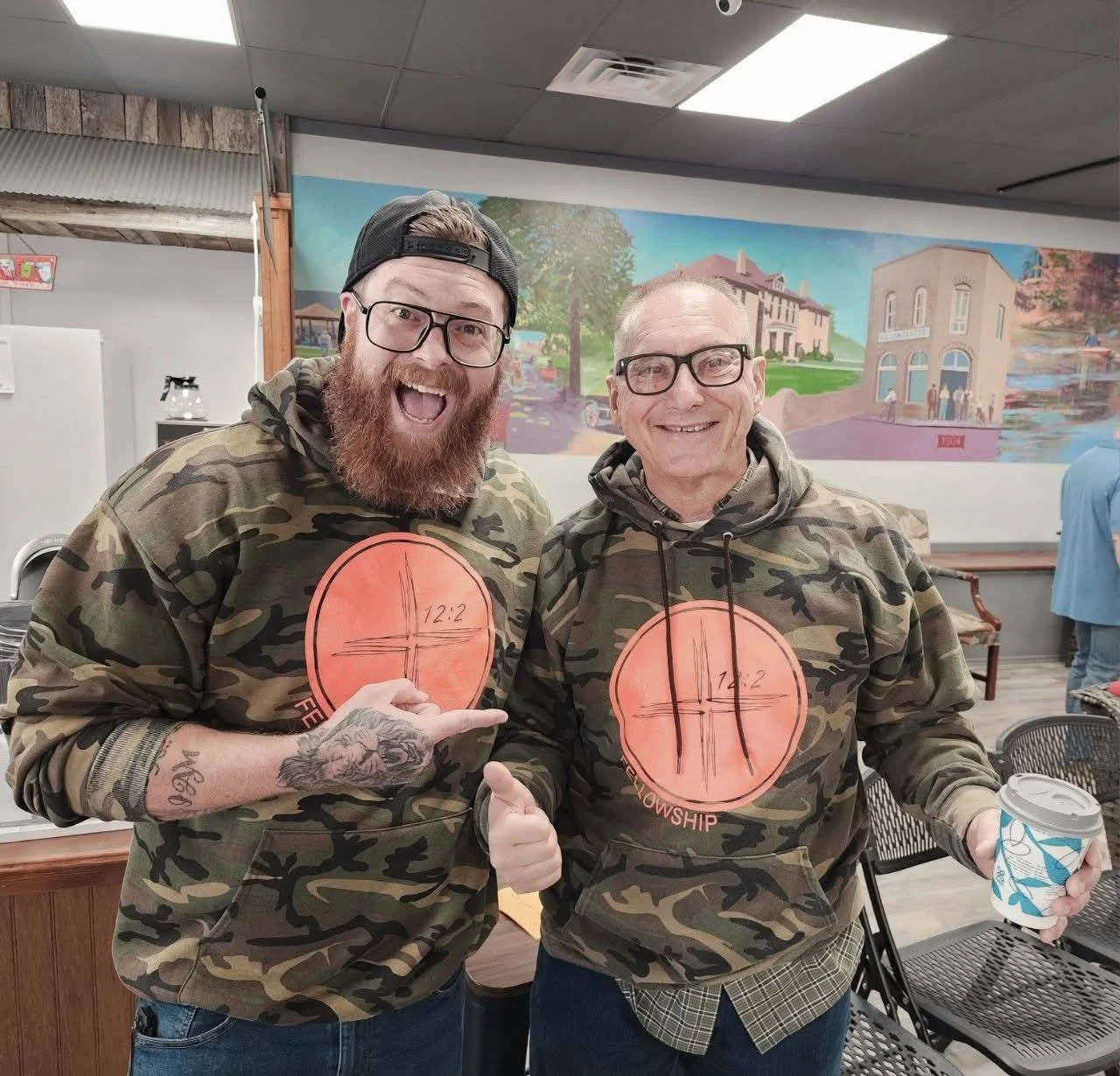 Two smiling men wearing camouflage hoodies with orange circular logos, one man pointing and the other holding a coffee cup inside a room with a mural on the wall.