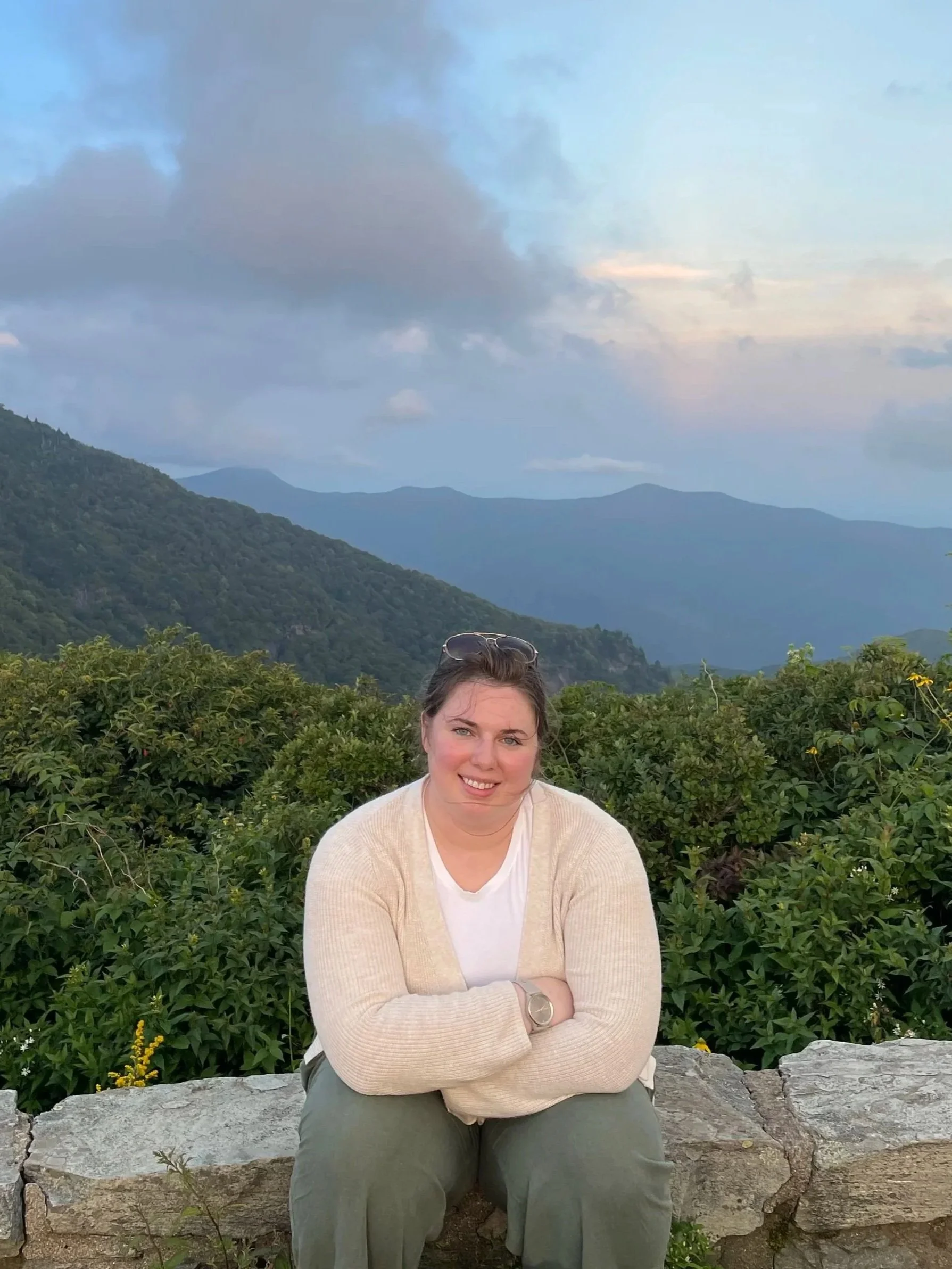 A young woman sitting on a stone wall in a mountainous outdoor setting during dusk, with mountains and clouds in the background.