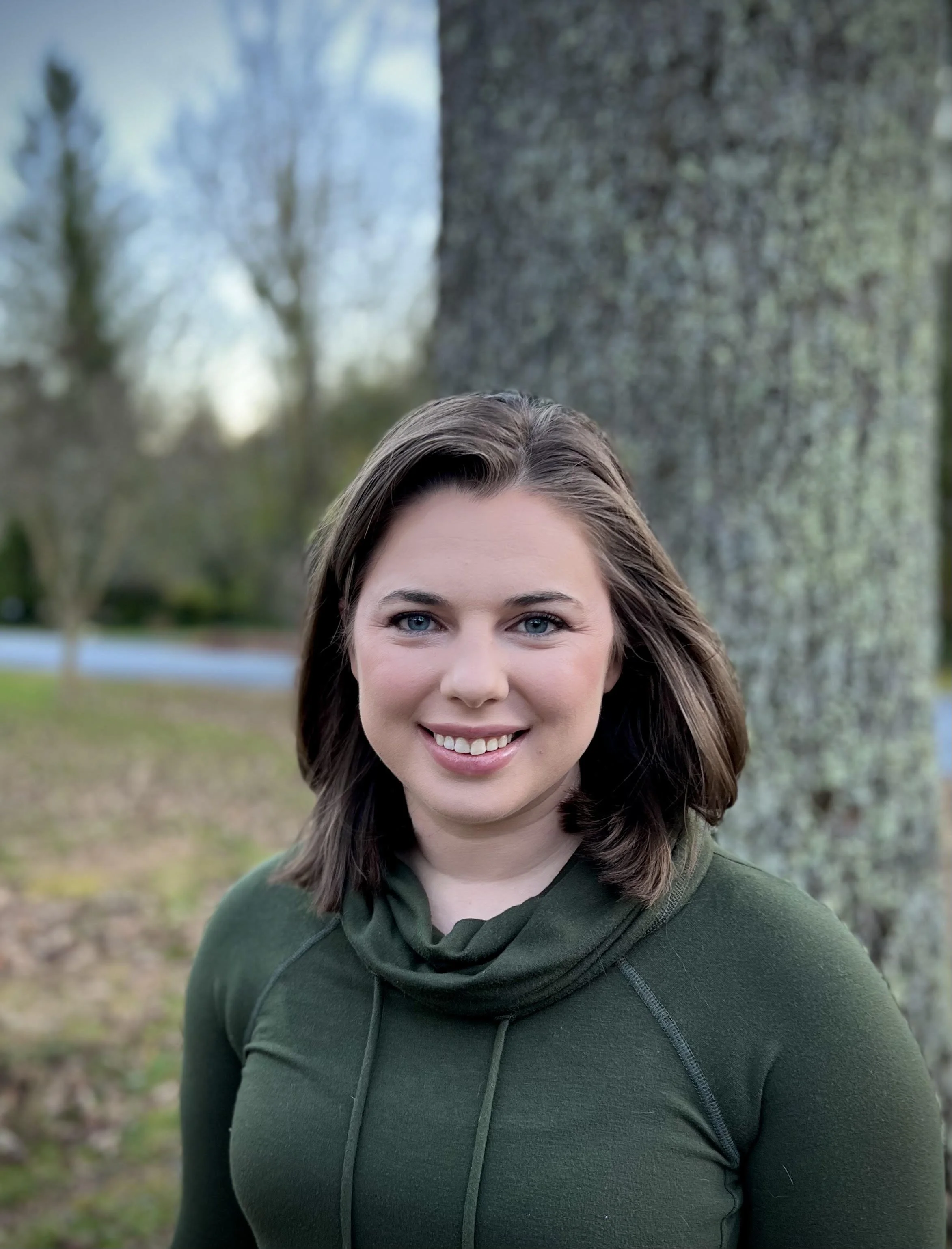 A smiling woman with shoulder-length brown hair and blue eyes, wearing a dark green top, standing outdoors in front of a tree with a blurred background of trees and a grassy area.