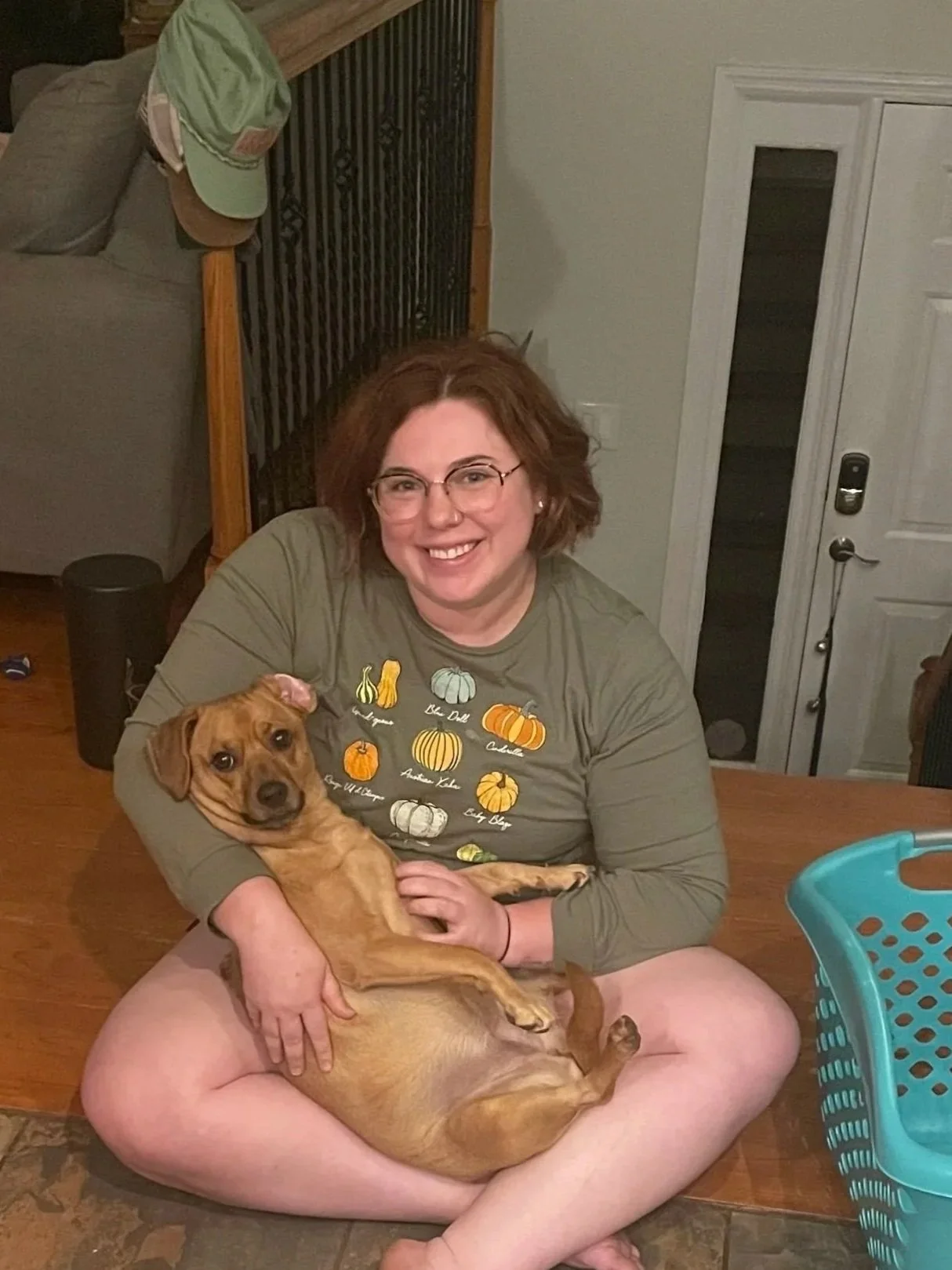 A woman with glasses and short, curly red hair sitting on the floor, smiling, holding a small brown dog with a relaxed expression. The woman is wearing a green long-sleeve shirt with pumpkin and squash illustrations and sitting cross-legged on a rug in a home setting.