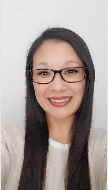 A woman with long dark hair wearing glasses and a white top, smiling in front of a white background.