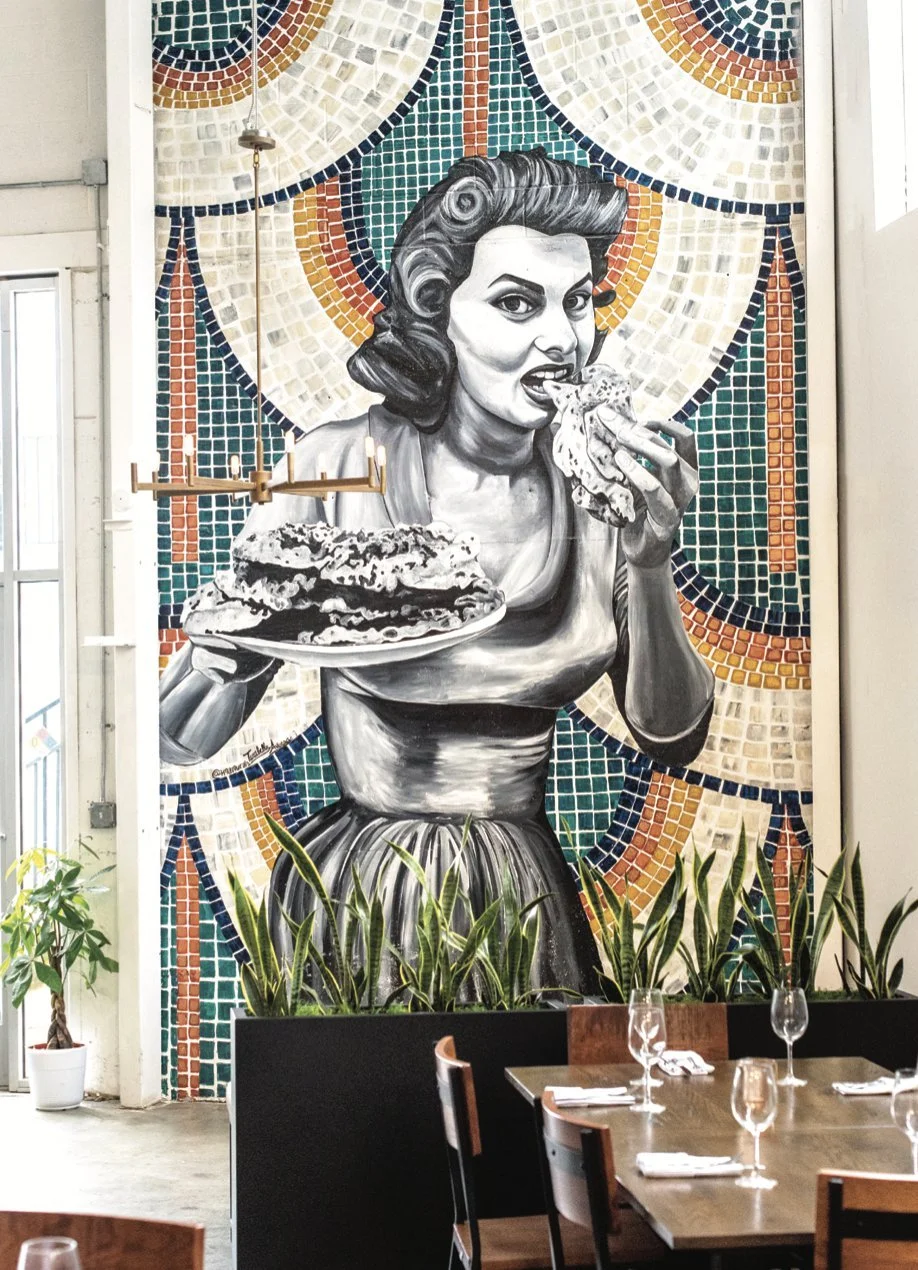 A large mural of a vintage woman with dark hair, eating pizza, standing against a colorful mosaic background. The mural is in black and white with a vibrant, intricate mosaic in the background. There are dining tables with glasses and napkins in the foreground, with some potted plants near the wall.
