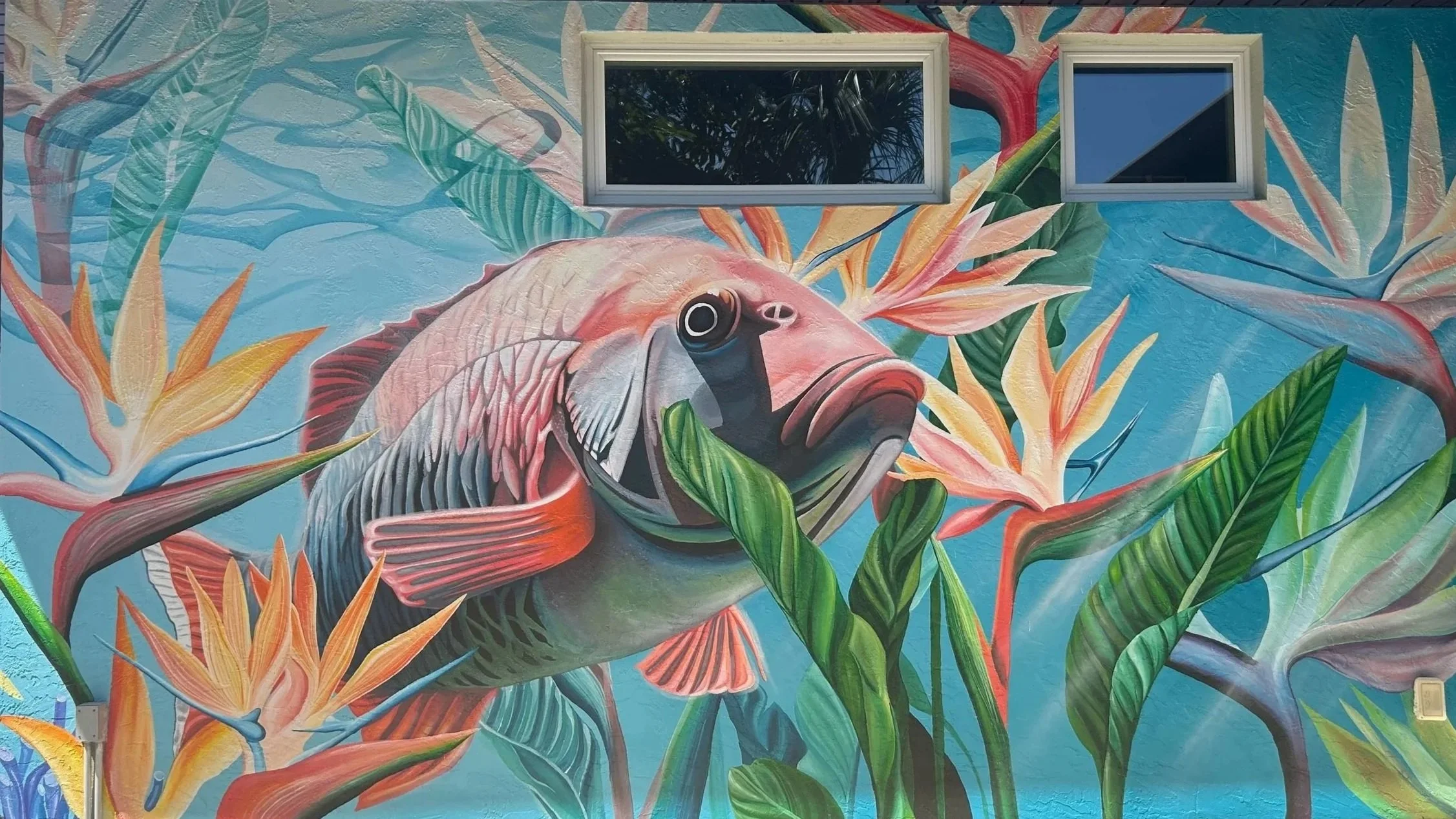 Colorful mural of a fish surrounded by tropical plants and flowers on a building wall, with two small windows at the top.
