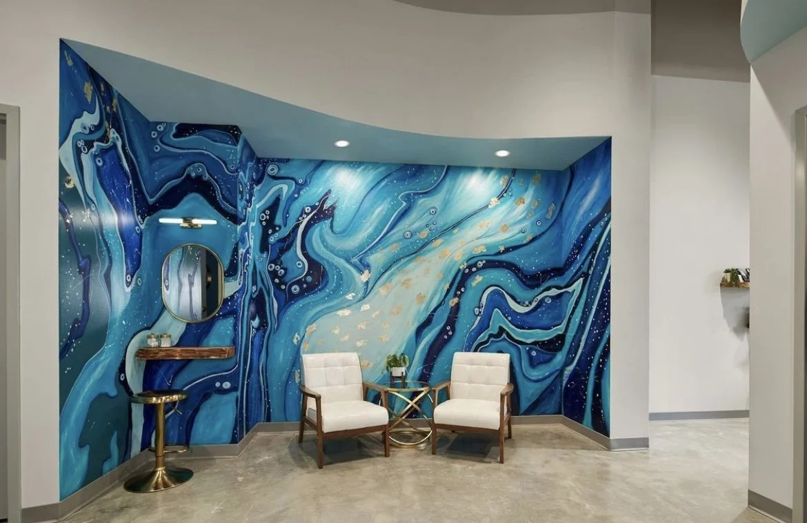 Indoor space with a vibrant blue abstract mural wall, two white armchairs, a small round gold table with a potted plant, and decorative shelves with small items.