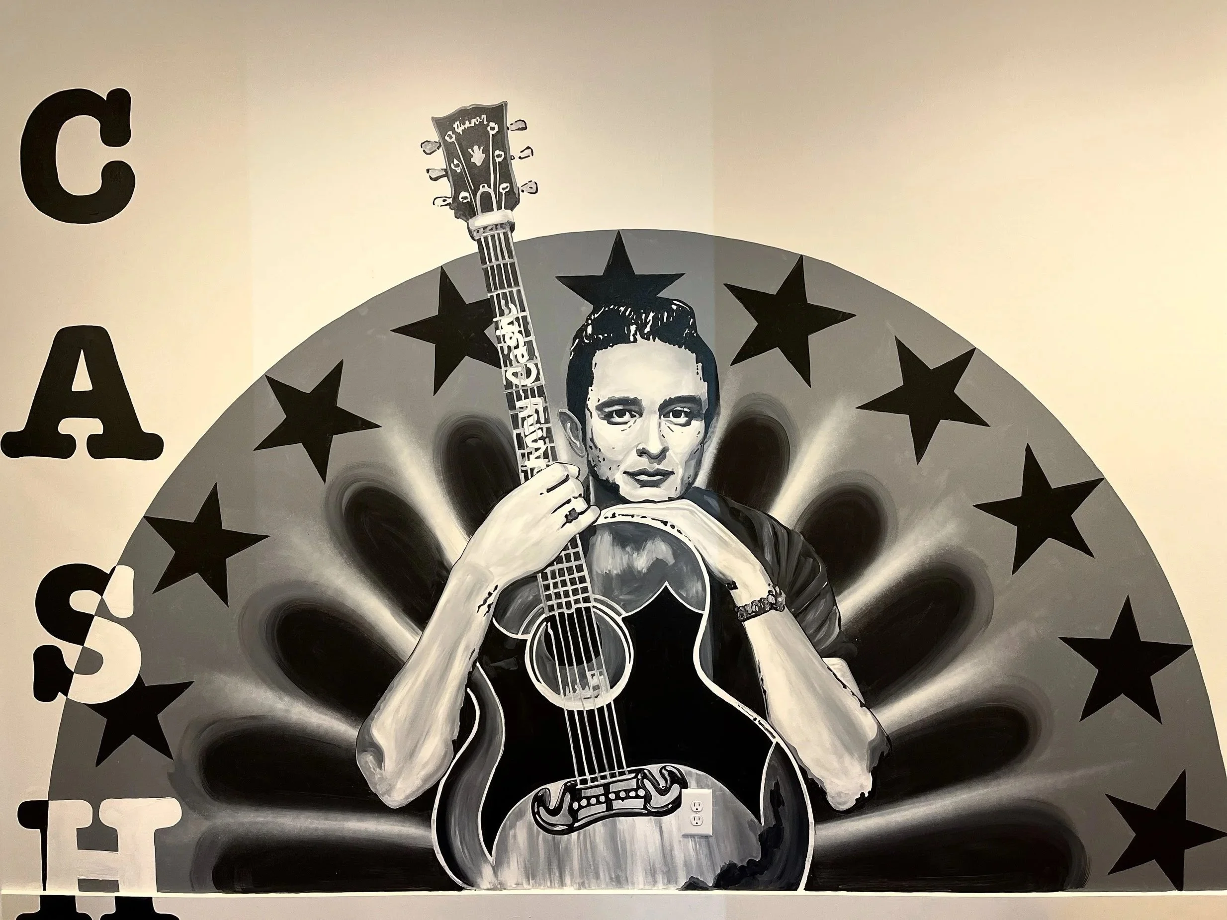 A mural of a woman with short dark hair holding an acoustic guitar, surrounded by a semi-circular design of black stars and black petal-like shapes, with large black letters spelling 'CAS' on the left.