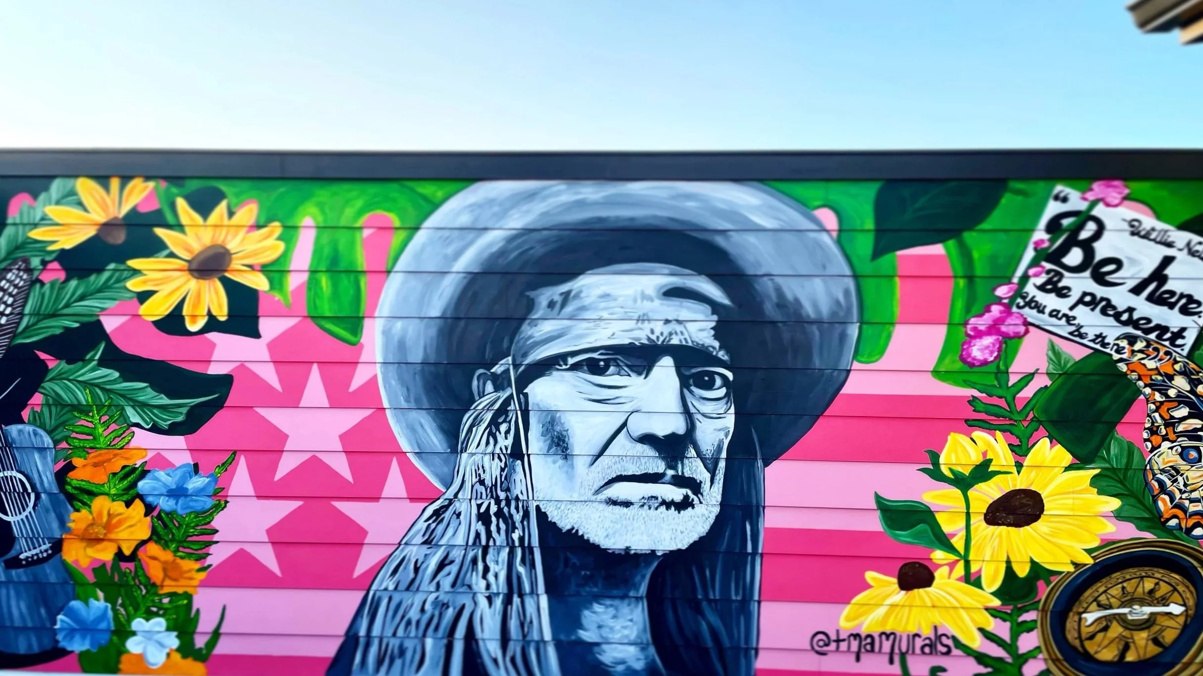 Colorful mural of an Indigenous elder wearing a large hat, surrounded by vibrant flowers, butterflies, and a quote that says 'Be here. Be present.'