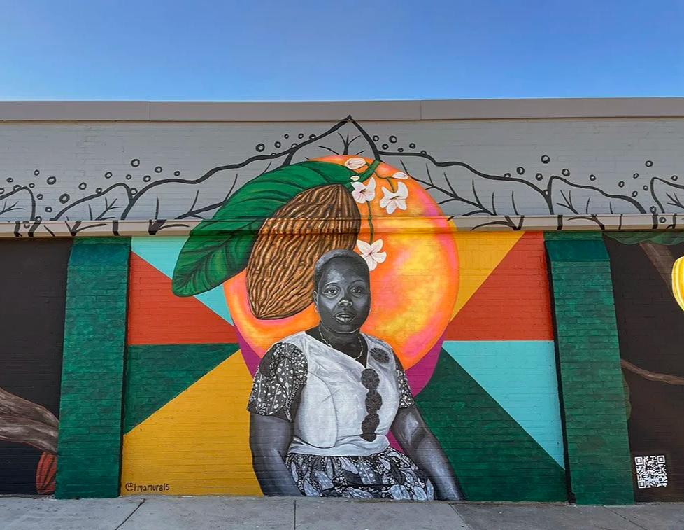 Street art mural of a young girl with dark skin and braided hair, painted in black and white, against a colorful background with geometric shapes and a large orange in the background. The mural also features a large almond and white flowers above the girl.