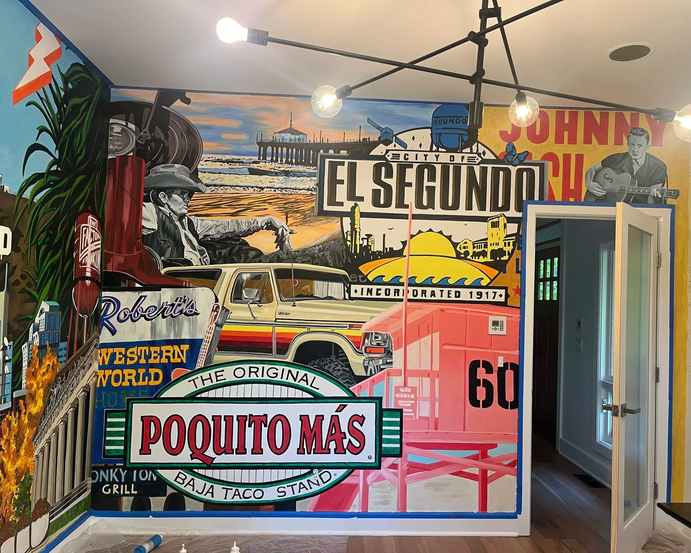 Colorful mural of a vintage roadside scene in El Segundo, California, featuring a large sign with the text "El Segundo", a classic pickup truck, a woman lying on the ground, and various retro and cityscape elements, including a pier, cars, and a landscape with sunset sky, all incorporating various local and cultural references.