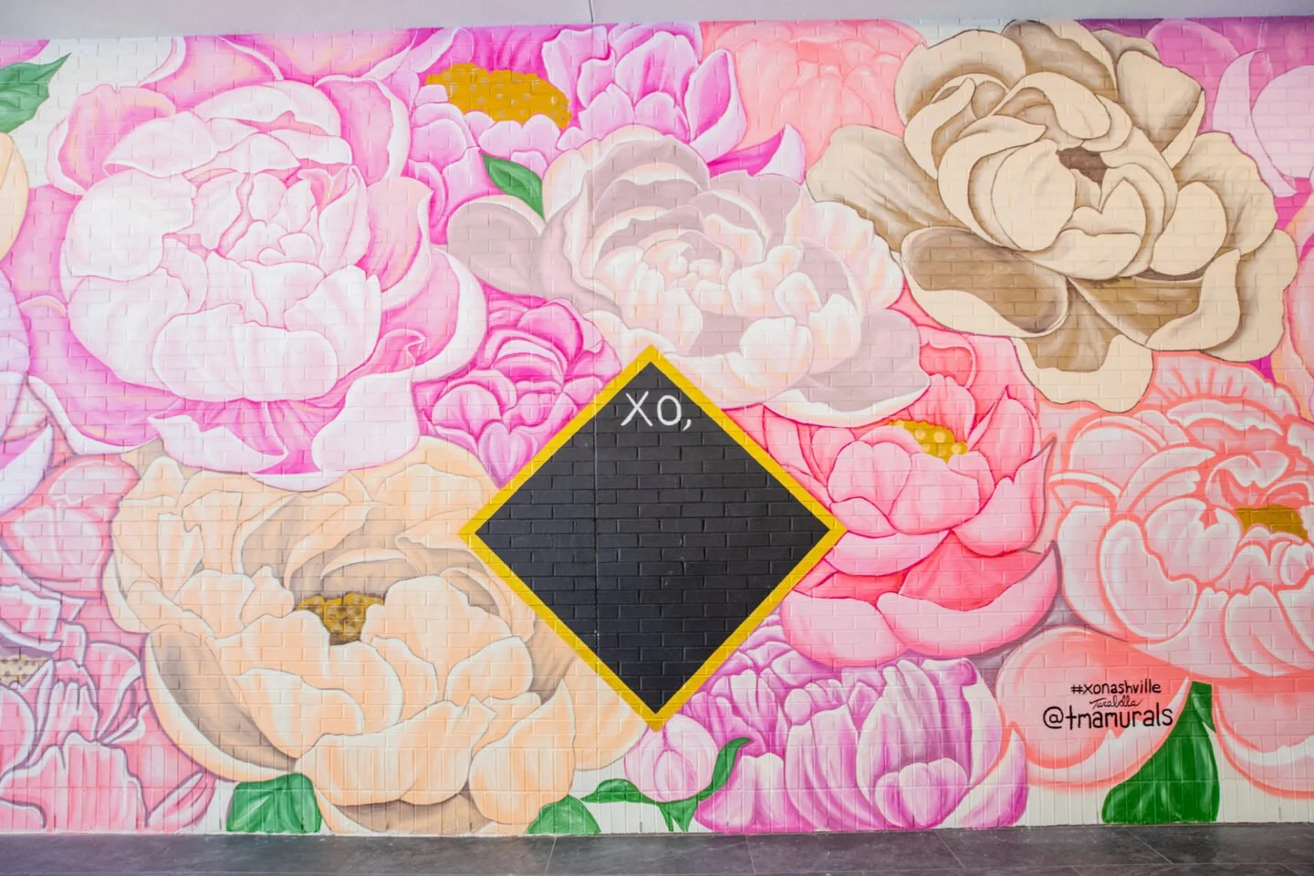 A colorful mural of large pink, white, beige, and peach peony flowers on a wall with the black XO symbol in the center, outlined in yellow.