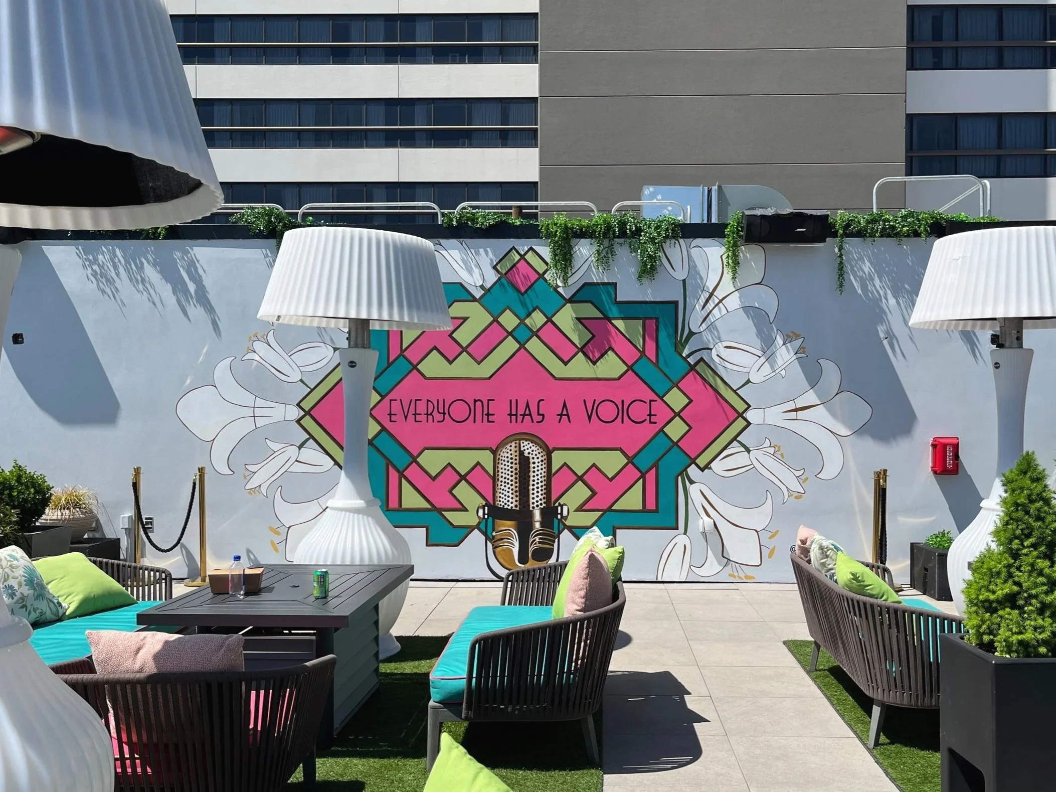 Outdoor patio with colorful mural reading 'Everyone has a voice', decorated with large white lamps, outdoor furniture, and potted plants.