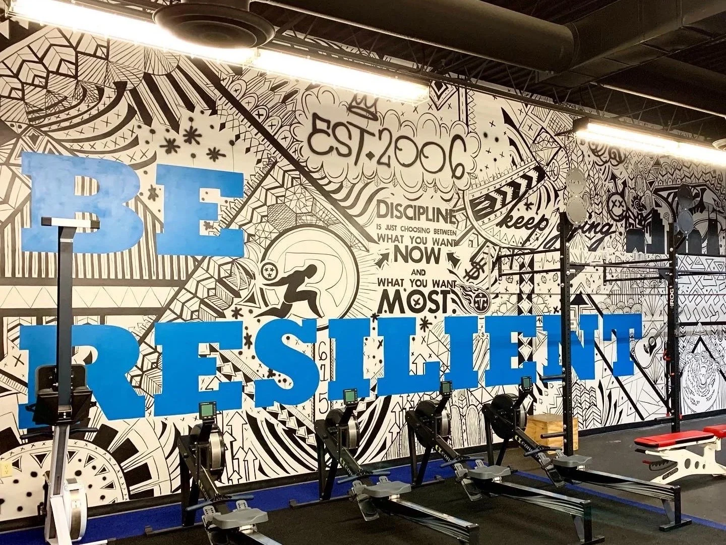 A gym interior with black and white mural art on the wall, large blue letters spelling 'BE RESILIENT', and rowing machines in the foreground.