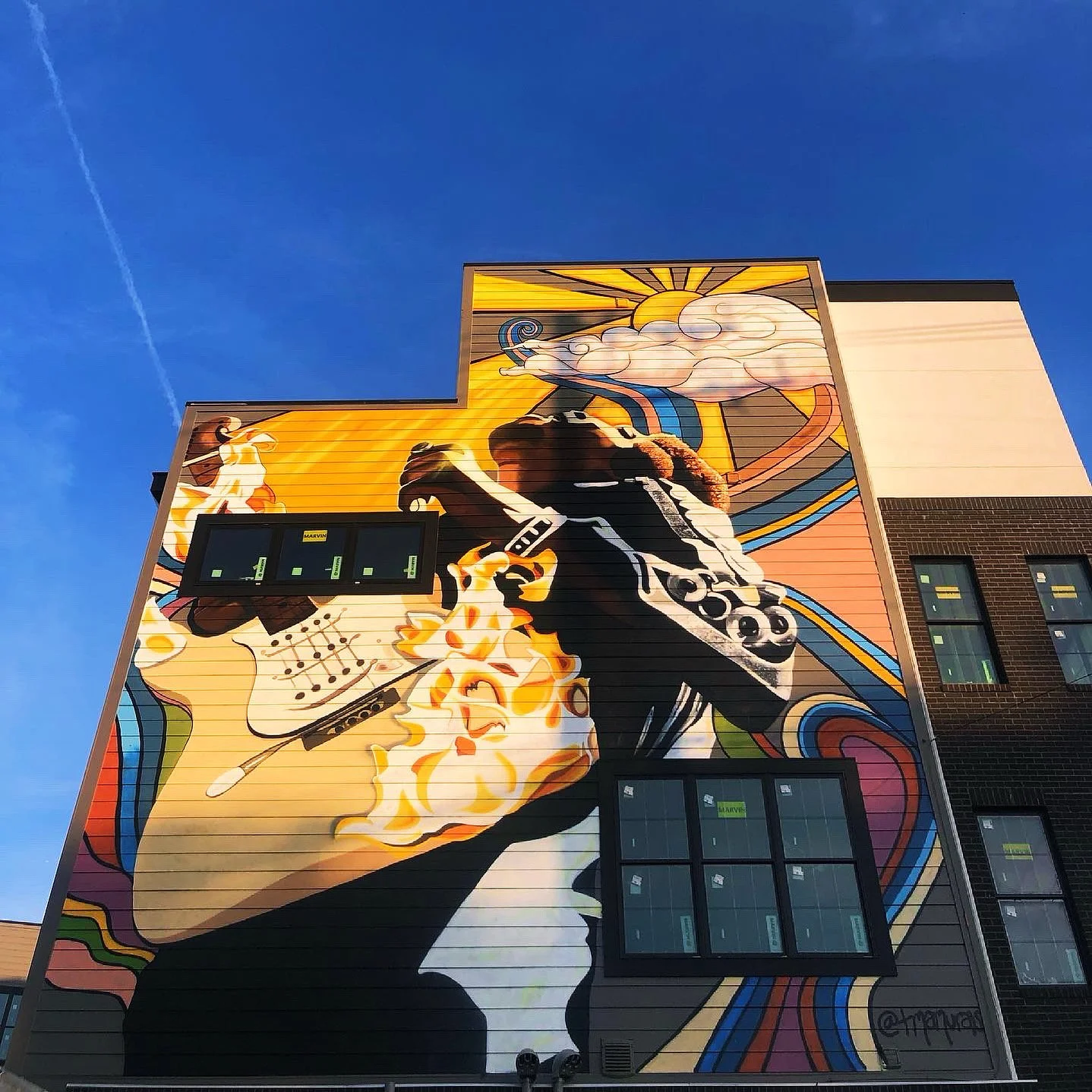 Colorful mural on the side of a building depicting a DJ playing music with abstract colorful shapes, a cloud with the sun, and a person dancing.