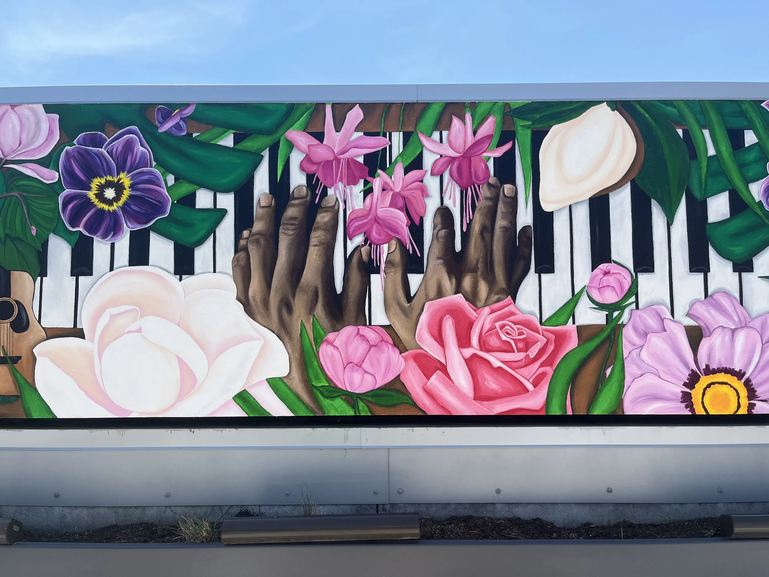 A colorful mural depicting two brown hands playing a piano with black and white keys. The piano is surrounded by vibrant flowers, including roses, orchids, and pansies, with a bright blue sky in the background.