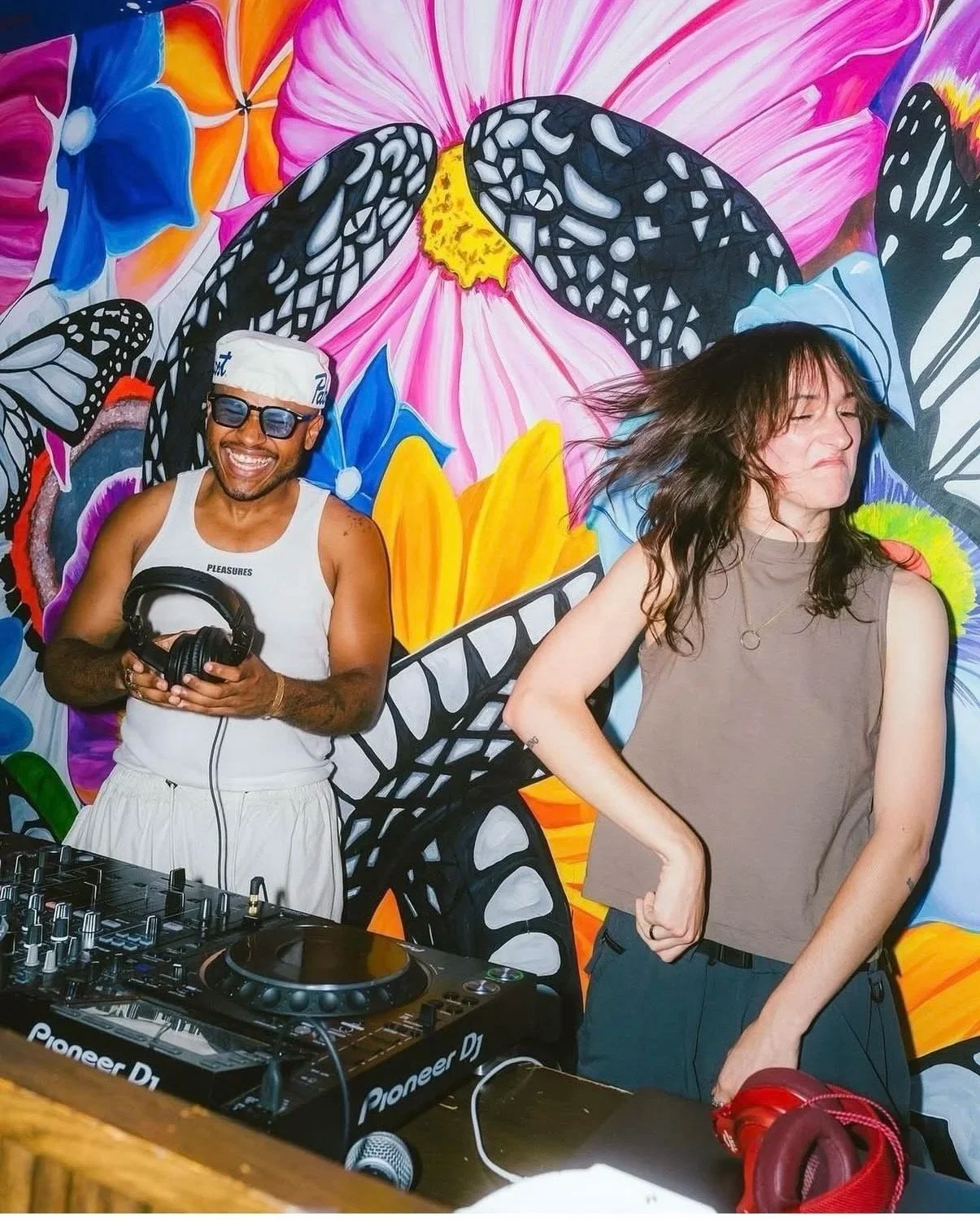 Two people dancing in front of a colorful butterfly mural. The man on the left is wearing sunglasses, a white tank top, a white head covering, and is holding headphones, smiling widely. The woman on the right has long dark hair, is wearing a sleeveless gray top, and appears to be dancing with her eyes closed.