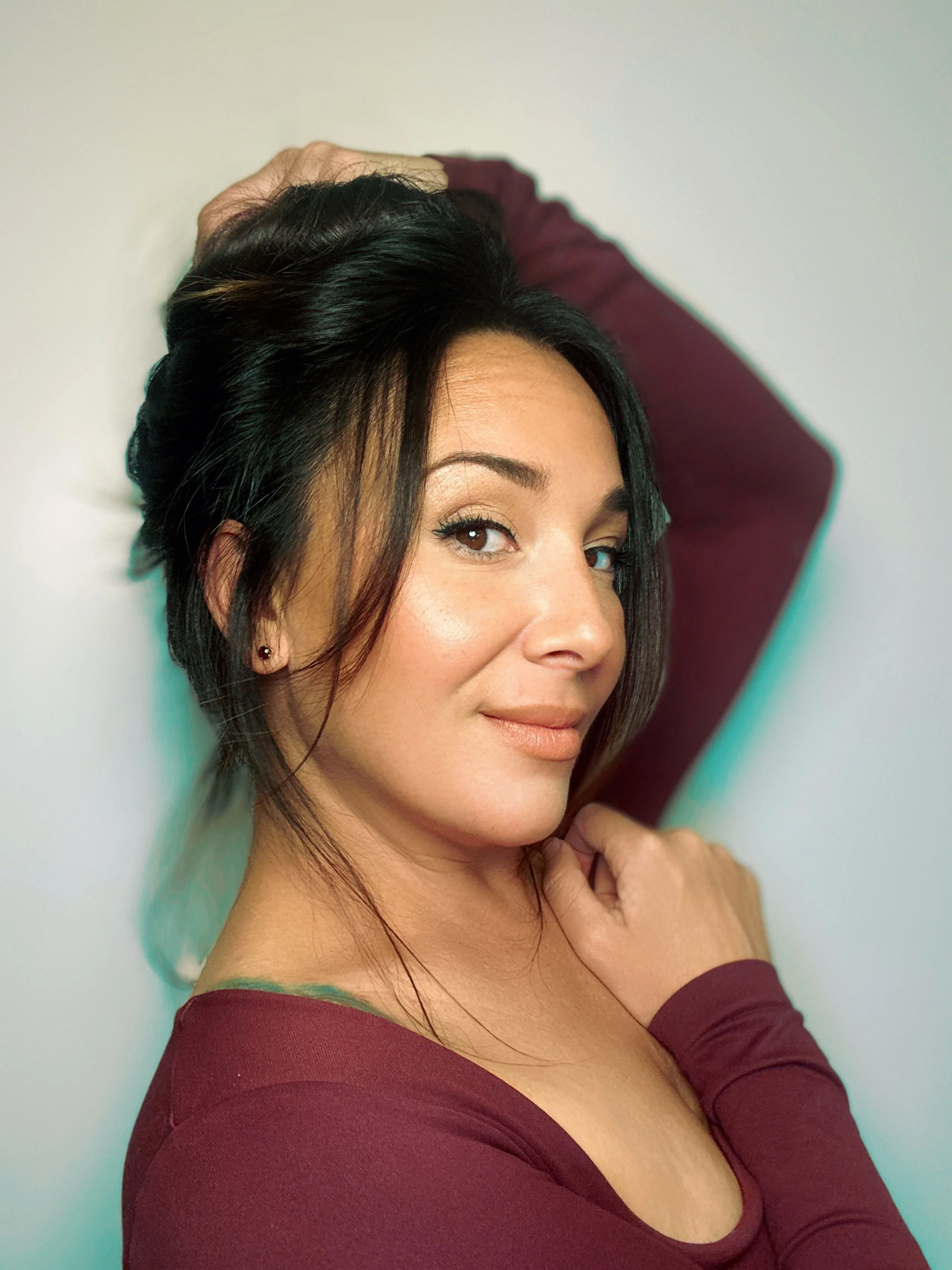 A woman with dark hair and makeup, wearing a maroon long-sleeve top, is posing with one arm behind her head and the other hand near her neck, against a plain light-colored background.
