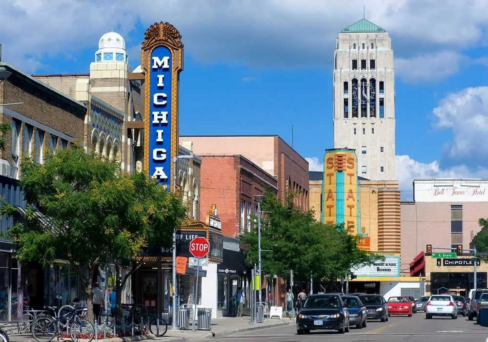 Downtown street scene with buildings, trees, cars, bicycles, a stop sign, and iconic signs for Michigan Theater and State Theatre in Ann Arbor, Michigan.